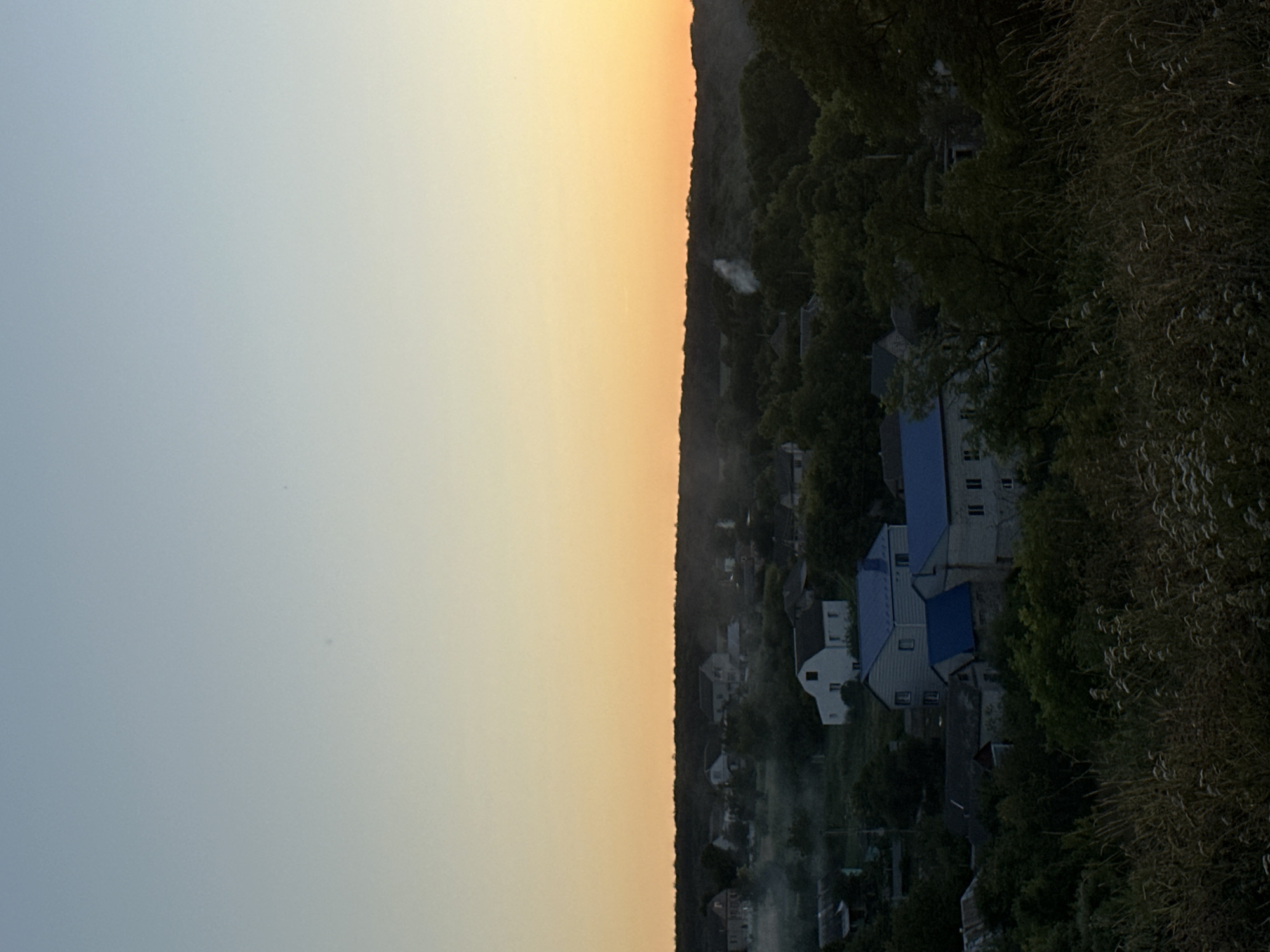 The image depicts a rural village scene at dusk, with a hazy horizon. In the foreground, tall, dry grass and small white wildflowers are visible. The middle ground shows several houses, some with blue roofs, nestled amongst dense green trees. Smoke is rising from a chimney in the distance. The background consists of a dark, treed hill. The sky transitions from a pale blue at the top to a warm orange near the horizon. This scene is located in the village of Makiv, Ukraine.