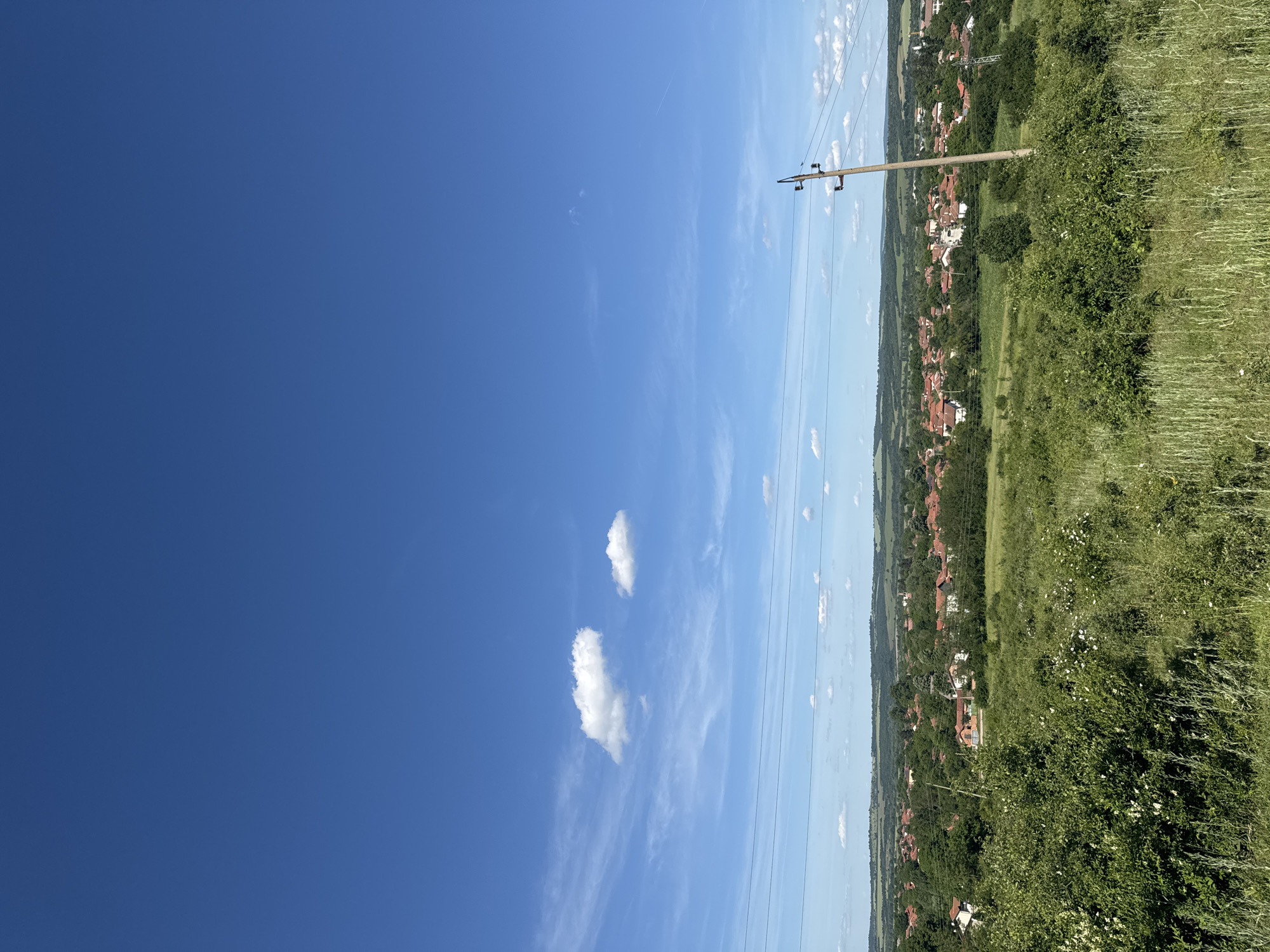 The image shows a landscape under a blue sky with scattered clouds. A power line pole is visible, running vertically through the scene. In the background, a settlement with red-roofed houses can be seen amidst green vegetation. The foreground consists of green grass and foliage. The image appears to be taken from a rural or semi-rural location. The city is not discernible.
