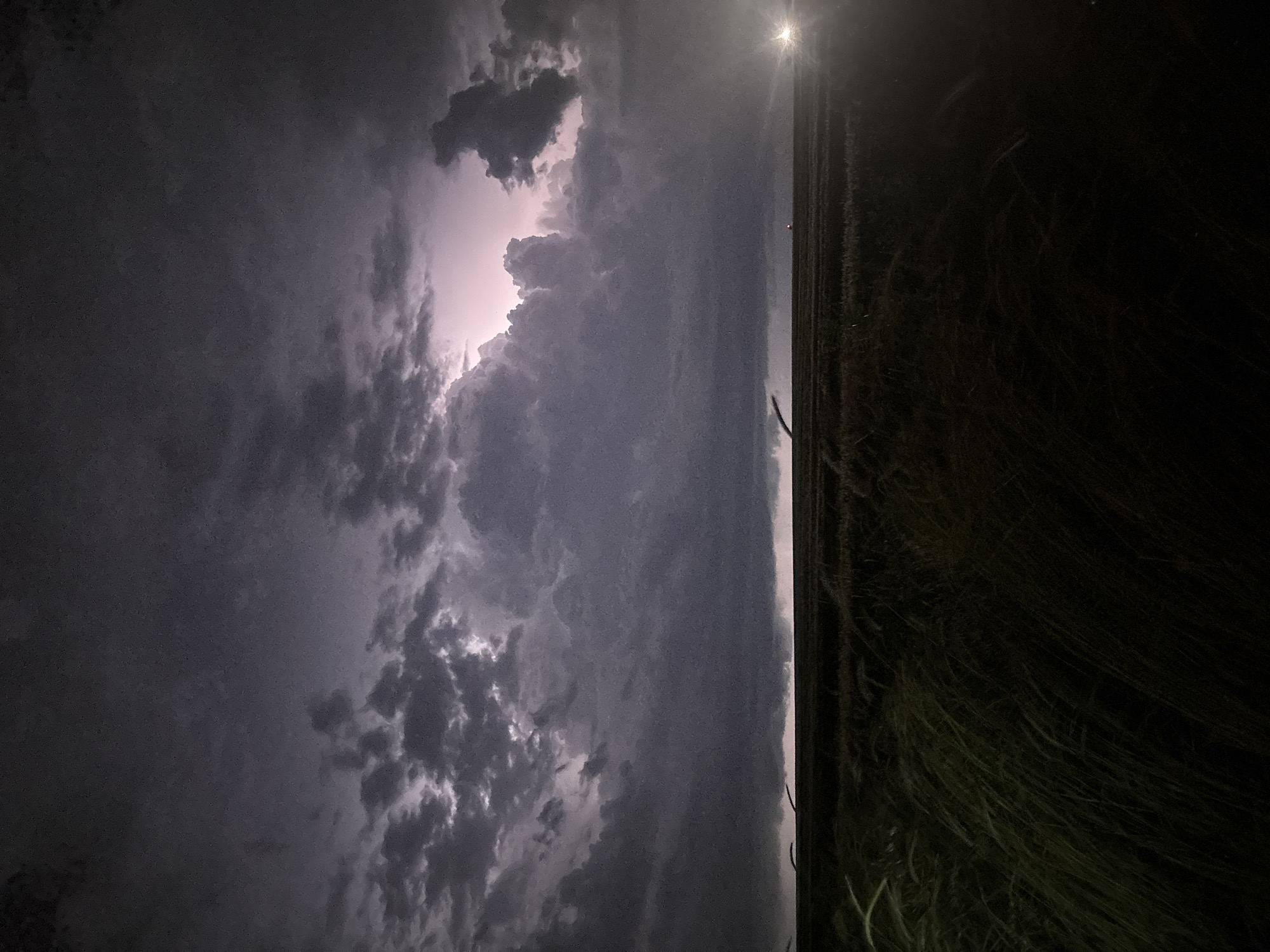 The image shows a night landscape, seemingly in a rural area. The sky is dominated by dark, stormy clouds with a bright flash of light, possibly lightning, illuminating the cloud formations. A bright light source, possibly the moon or a strong artificial light, is visible above the horizon. The foreground consists of a field of tall grass or vegetation that is dark due to the lack of light. The photograph is oriented vertically. There is no evidence to suggest this image was taken in Burgas, Bulgaria. There are no people present in the image.