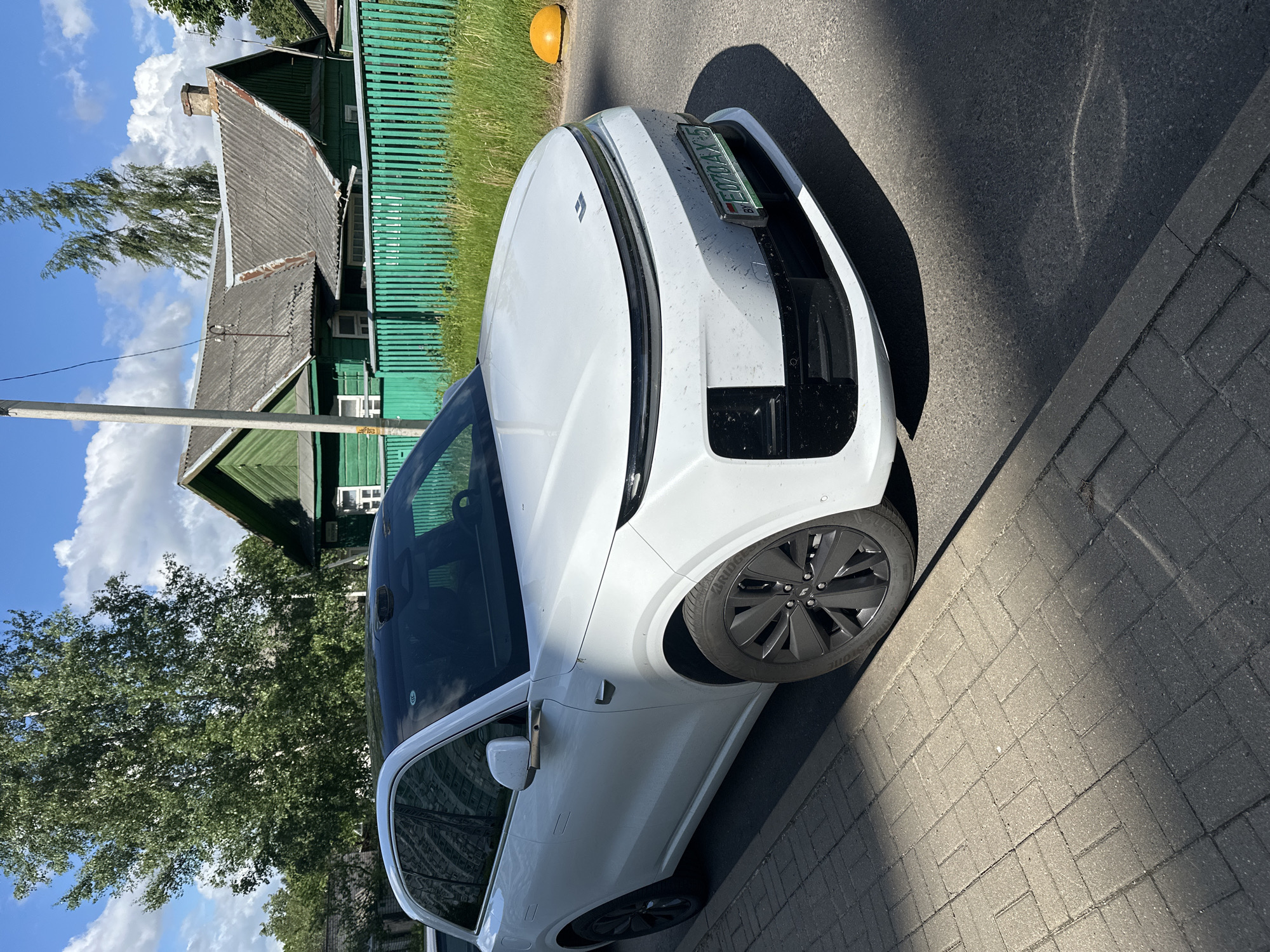The image depicts a white electric car parked on a paved street in what appears to be a residential area. A traditional wooden house with green siding and a gray roof is visible in the background. The car has a license plate. The image was taken in Minsk, Belarus.