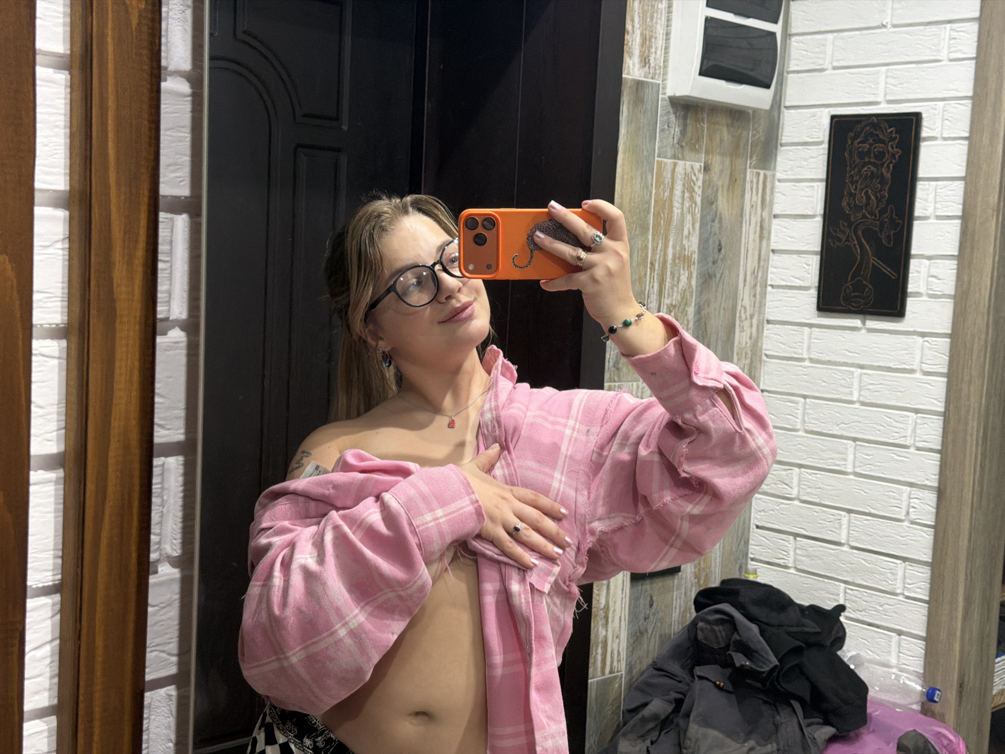 A young woman is captured taking a mirror selfie in an indoor setting, likely a home in Большая Шильна, Russia. She has light brown hair and wears black-rimmed glasses, looking towards her phone screen with a calm, slightly smiling expression. She is dressed in an open pink and white plaid long-sleeved shirt, revealing her midriff. Her right hand holds an orange-cased smartphone, which features a distinctive leopard design on its back, while her left hand rests on her chest, holding the shirt.
She is adorned with earrings, a silver necklace with a small red charm, a beaded bracelet with green and black beads, and multiple rings on her fingers. Her fingernails are painted a light, possibly white or silver, color. A white clothing tag is visible on the left shoulder of her shirt, but any text on it is illegible.
The background displays diverse textures, characteristic of a residential interior. To the left, there is a dark paneled door, followed by vertical wooden panels and a white brick-patterned wall. On the right, a textured grey and brown tile-like wall is visible, along with a light wooden trim. A black rectangular plaque, adorned with an ornate bearded figure, is mounted on the white brick wall. In the lower right corner, a stack of dark clothing, including jackets, is piled up, next to a plastic bottle with an unreadable blue label on its cap. The lighting suggests an indoor environment, likely illuminated by artificial sources.