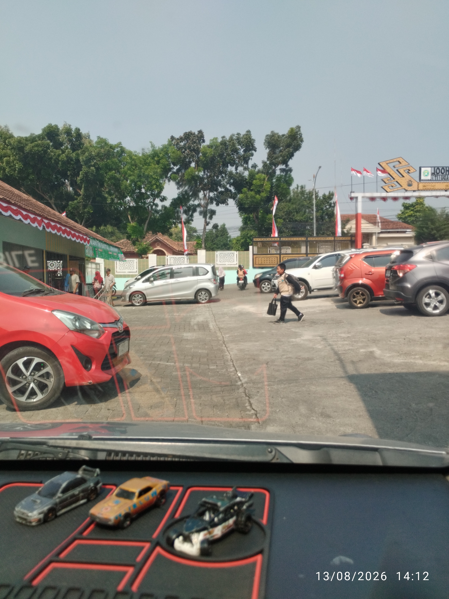 The image depicts a street scene in Bandar Lampung, Indonesia, on August 13, 2026, at 2:12 PM. In the foreground, the dashboard of a vehicle is visible, displaying three toy cars. The scene transitions to an outdoor area with several parked cars, including a red car in the left foreground, a silver car in the middle ground, and an orange and a dark gray car further back. A young male wearing a backpack walks across the paved area, carrying a black bag. In the background, a building with a red and white awning is present, along with trees and flags, suggesting a public or institutional setting. The sky is clear and bright.