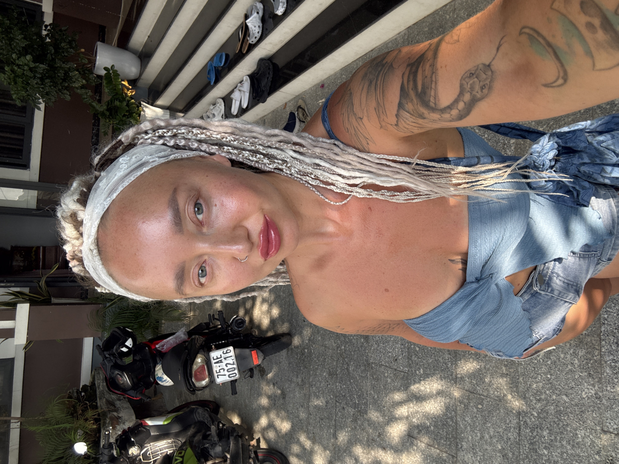 The image shows a woman with blonde braided hair, wearing a blue top and denim shorts, posing for a selfie outdoors. She has a snake tattoo on her right arm. In the background, there are two scooters parked on a paved area. Further back, there are stairs leading up to a building with plants and white pots. The scene appears to be in Bac Nha Trang Ward, Vietnam, as indicated by the license plate on one of the scooters which reads "75-AE 002.16". The lighting suggests it is daytime, possibly late afternoon, with dappled sunlight filtering through foliage. Several pairs of shoes are visible on the stairs.