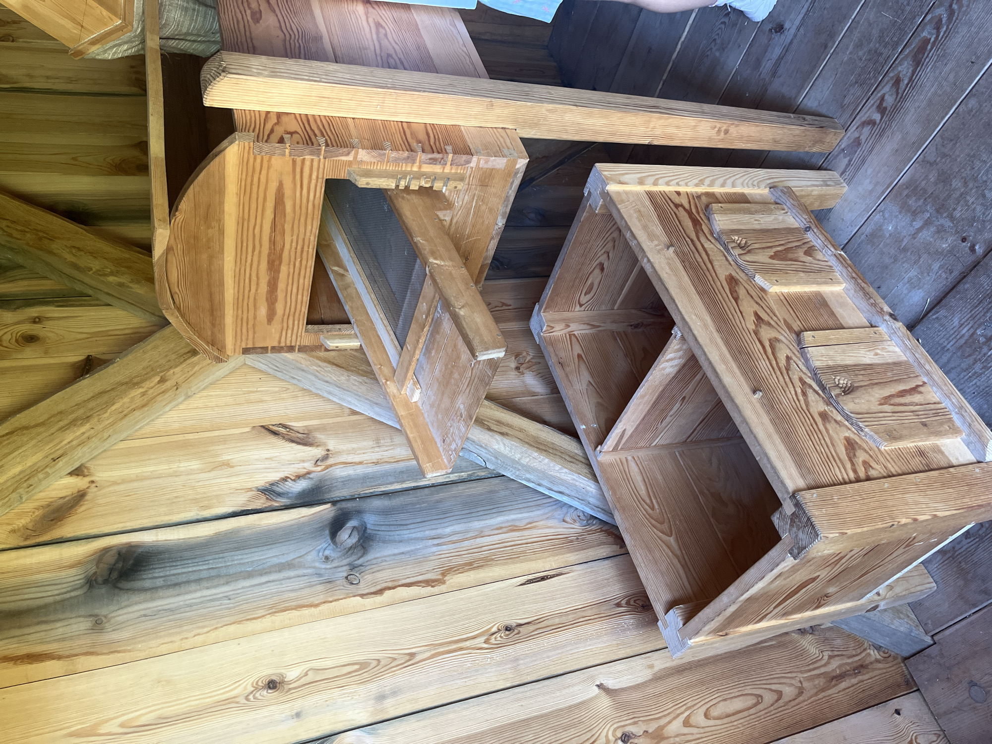 The image shows a close-up of a wooden machine, possibly a grain sorter or a similar agricultural device, located indoors in what appears to be a rustic building. The machine is constructed from light-colored wood, with visible grain patterns and some older, darker wood integrated into the structure, likely part of the building's wall or support beams. The machine itself features a hopper at the top with a sieve or screen, from which material would presumably fall into a collection bin below. The bin is also made of wood and is divided into sections. The floor is also made of wooden planks. In the lower right corner, the legs of a person are visible, wearing white shoes and blue shorts, suggesting that this is a functional display or a working area. Based on the architectural style and the likely historical context of such a machine, this scene is consistent with a display at the Kuressaare Castle Museum in Kuressaare, Estonia, which often features historical agricultural equipment.