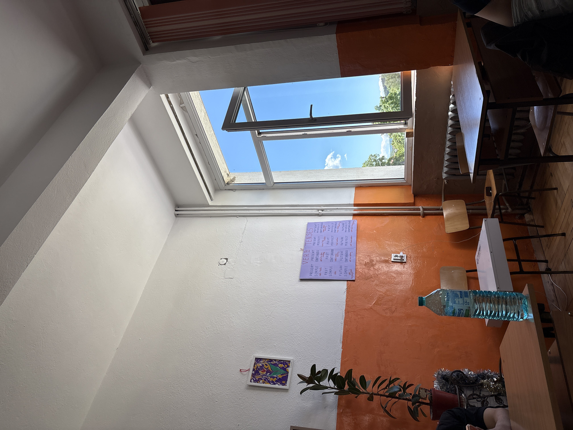 The image shows a classroom interior with a tilted camera angle. A large skylight is open, revealing a blue sky with some trees visible outside. The walls are painted with a split design, white on the left and orange on the right. A "VERB TENSES" poster is on the wall. Below it is a small framed piece of artwork and a potted plant. A water bottle is sitting on a desk, with school chairs and desks visible. There is a row of tables, some of them occupied by people. The scene appears to be in a classroom setting. There is no information about the location in the city of Panagyurishte, Bulgaria.