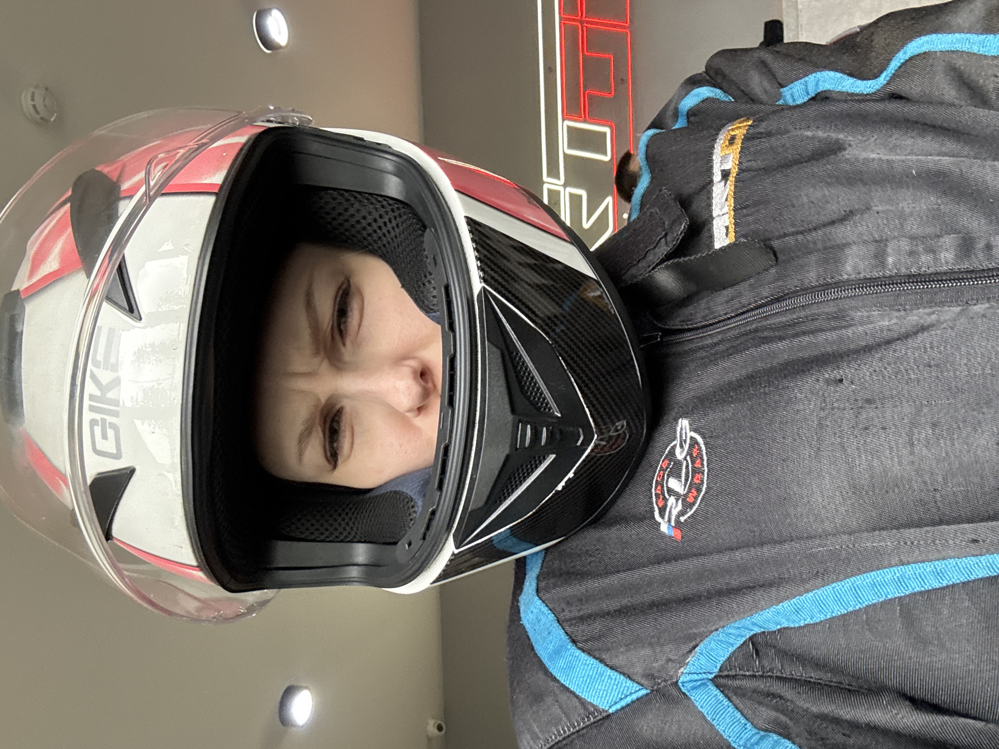 A woman is wearing a full-face helmet with a transparent visor. The helmet is primarily white with red accents and the letters "GIKE" visible. Underneath the helmet, a blue face mask is partially visible. She is also wearing a black racing suit with light blue trim and a circular logo with white, red, and black details. The background appears to be indoors with a neutral-toned wall and a blurred neon sign. Based on the logo on the racing suit, this image was taken at the Perm Motorsport Complex in the city of Perm, Russia.