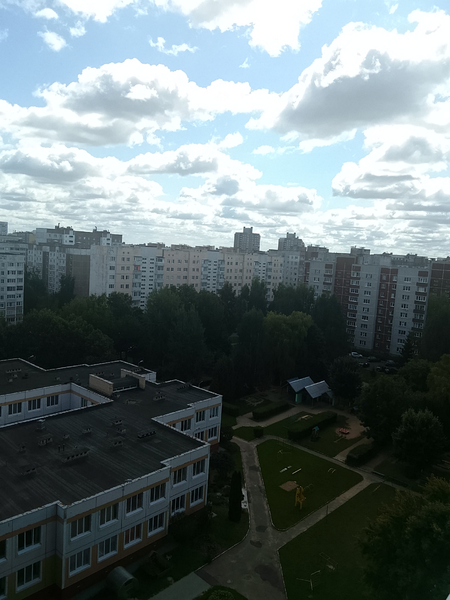 The image depicts an aerial view of a residential area in Minsk, Belarus, on a cloudy day. In the foreground, two school buildings with dark, flat roofs are visible. The buildings are multi-story with rows of windows. Beyond the schools, a dense canopy of green trees separates the foreground from a series of apartment buildings in the mid-ground. These apartment buildings are tall and vary in architectural style and color, with many windows and balconies. Some buildings appear to be older Soviet-era constructions, while others show more modern designs. In the background, the cityscape extends with more buildings and a hazy sky filled with white and gray clouds. A park or recreational area with playground equipment and a yellow giraffe-shaped structure is situated between the trees and the apartment buildings. A few cars are parked on a road near the apartment buildings. The overall impression is one of an urban residential district with a mix of older and newer constructions, surrounded by trees and under a dynamic cloud cover.