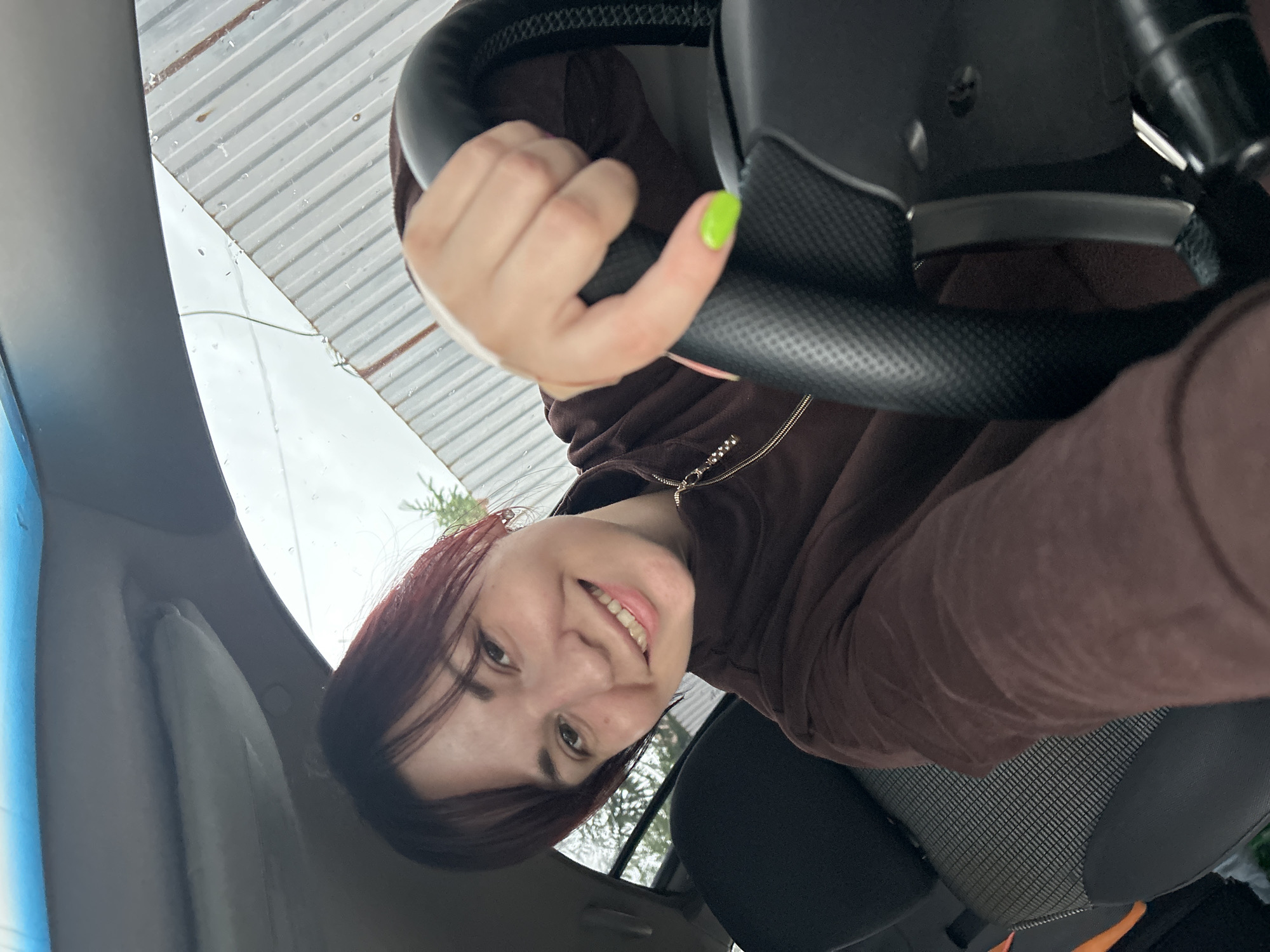 A young woman with dark hair, wearing a brown velour zip-up jacket, is seated in the driver's seat of a car. She is smiling and looking at the camera, with her right hand resting on the steering wheel. The steering wheel is black and textured. The interior of the car is visible, including the seat fabric and part of the dashboard. Outside the car, a corrugated metal fence and some trees are visible. The weather appears to be overcast with some rain. The scene is likely located in the city of Fokino, Russia.
