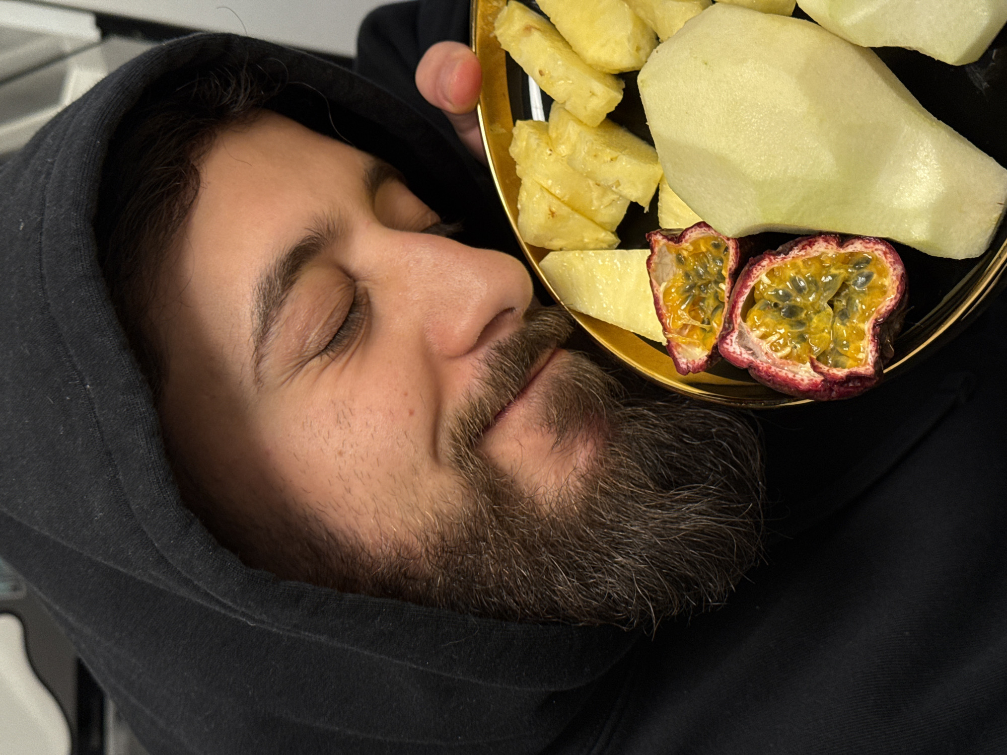 A close-up image displays an adult male, seen from the upper torso, wearing a black hooded sweatshirt with the hood up. His head is angled down towards a round, dark-colored plate with a golden rim, which he holds with his right hand (thumb and forefinger visible). His eyes are closed, and a slight smile is visible, suggesting he is smelling the contents of the plate. He has a dark brown beard and mustache. The plate contains an assortment of cut fresh fruit: multiple yellow, rectangular pieces of pineapple, two halves of a passion fruit displaying orange pulp and black seeds, and several irregularly shaped pieces of peeled, pale green or yellow fruit, possibly pears or apples. The background is an indistinct, light-colored, out-of-focus indoor environment. This scene is located in the city of Strasbourg, France.