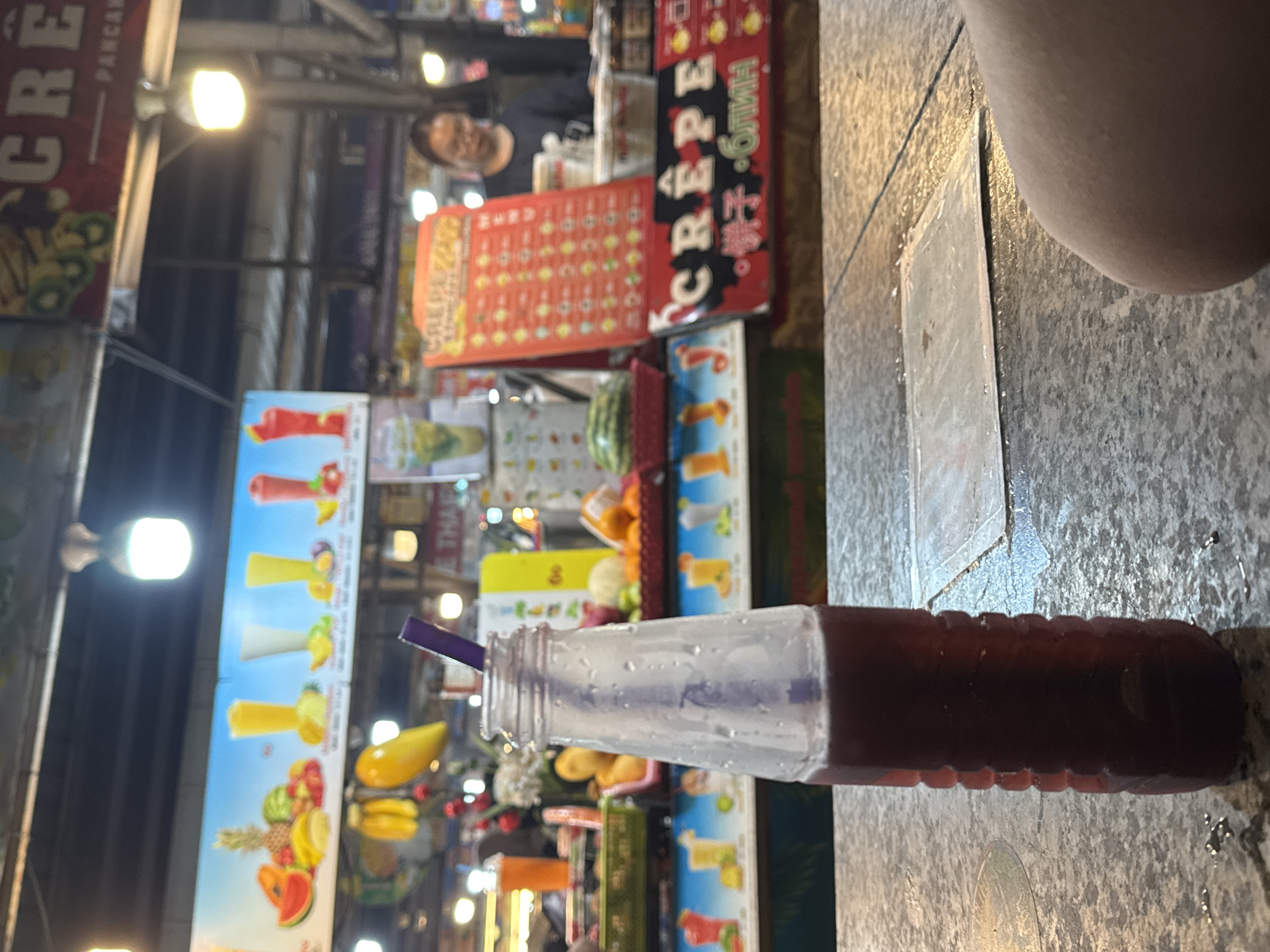 The image depicts a street food vendor setting in Patong, Thailand. A red beverage in a plastic bottle with a purple straw rests on a gray, possibly stone, surface. In the background, a man stands behind a food stall displaying menus with images of crepes, pancakes, and fruit smoothies. The stall is adorned with colorful signs written in both English and Thai. Streetlights provide illumination, and there is a sense of bustling nightlife due to the background activity.