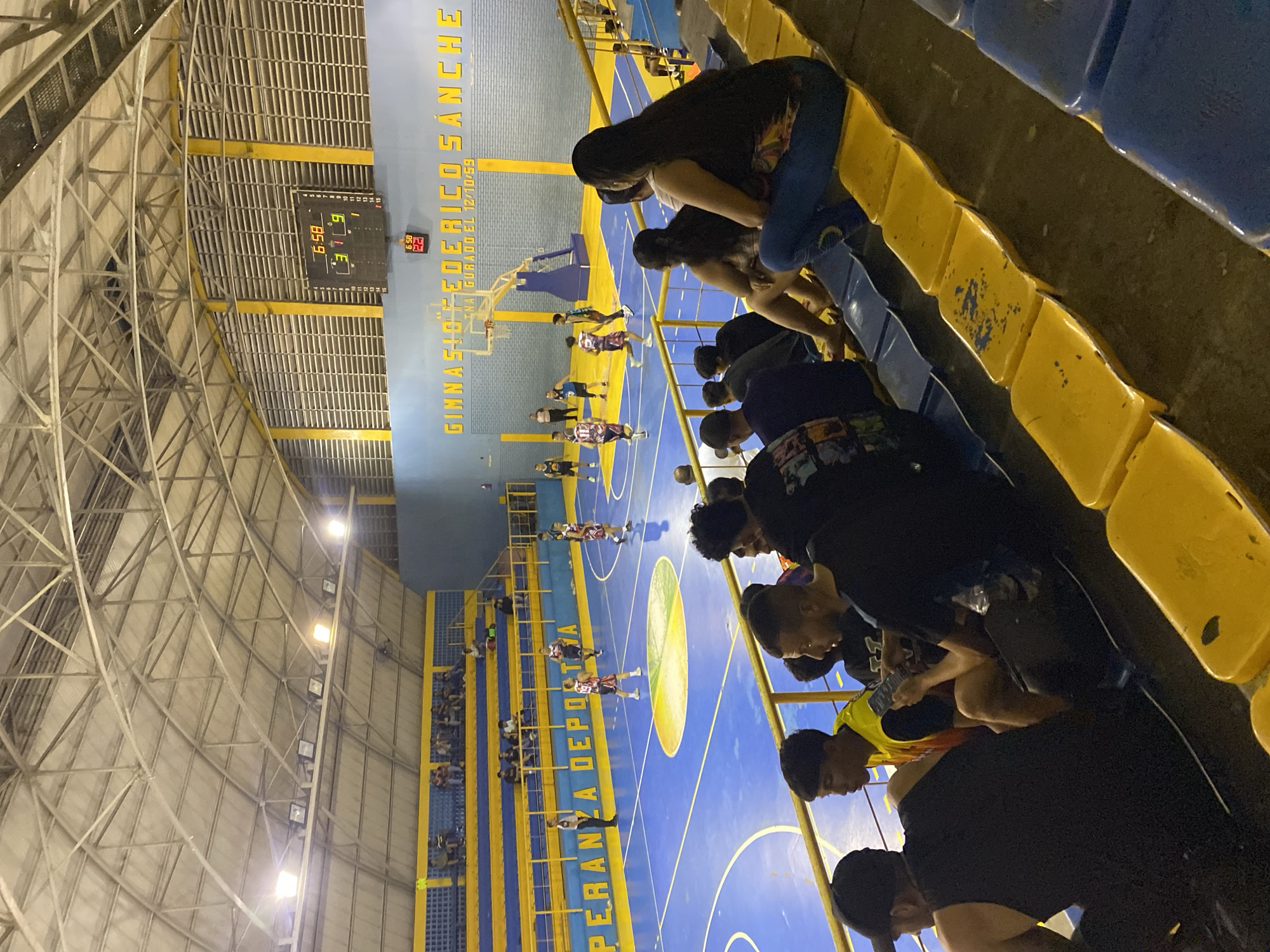 The image shows an indoor basketball game in progress at Gimnasio Federíco Sánchez, in Tinaquillo, Venezuela. Spectators are seated in blue and yellow stadium-style seats on one side of the court. Players are visible on the blue and yellow basketball court. The scoreboard shows a score of 31 to 22 and a remaining time of 6:58. The gym has a large, domed ceiling with a metal support structure.