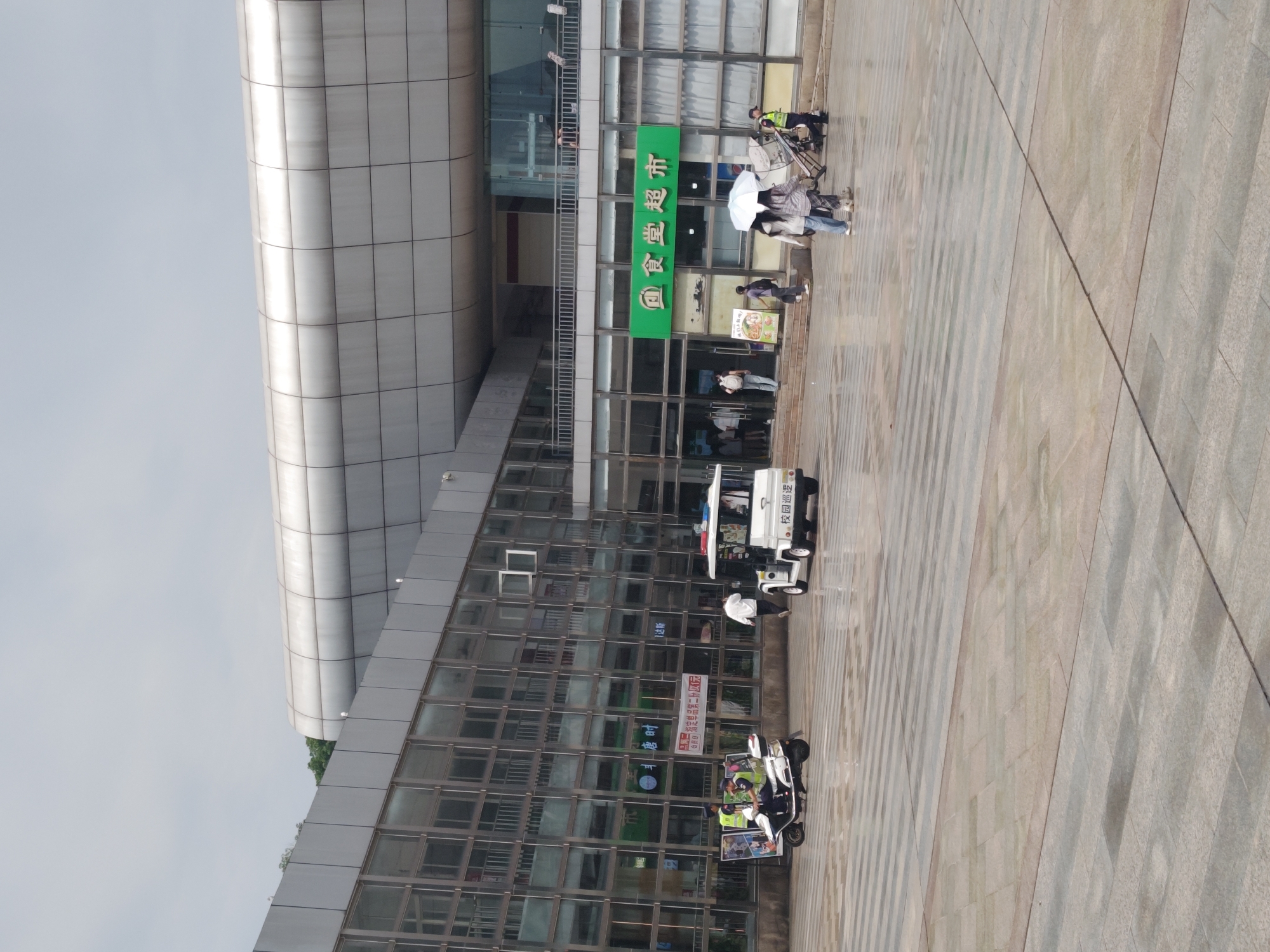The image depicts an outdoor scene in front of a building with a modern, metallic facade. The building features large, reflective glass windows. In the foreground, the ground is paved with large, rectangular tiles arranged in a patterned walkway.
Several people are present in the scene. On the left, two individuals wearing yellow vests and helmets are seated on a white three-wheeled electric vehicle. One person is driving, and the other is standing. To the right of this vehicle, a white electric vehicle with flashing lights, resembling a security or patrol car, is parked. A person is walking away from it towards the building entrance.
In the middle of the scene, a group of people are walking on the paved area. One individual is holding a white umbrella, and another person is pushing a stroller. A security guard in a yellow vest is positioned near the stroller. Further back, several people are entering and exiting the building through glass doors.
Above the entrance to the building, a large green sign with white Chinese characters reads "食堂超市" (Shitang Chaoshi), which translates to "Canteen Supermarket". The overall environment suggests a public space, possibly near an educational institution or a commercial complex. The sky is overcast.
Based on the provided information, this image was captured in the city of Chongqing High-tech Zone, China.
