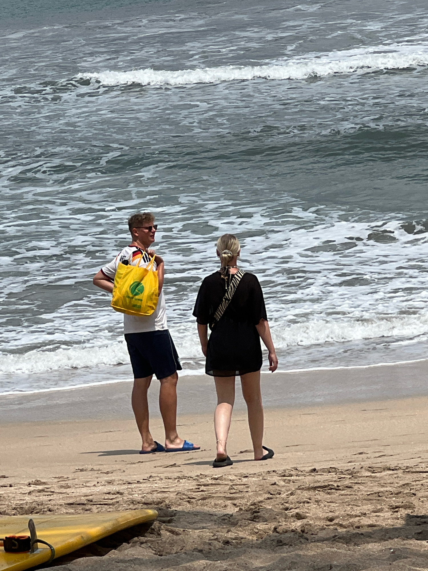 Two individuals stand on a sandy beach, facing the ocean in Kuta, Indonesia. The man on the left, wearing sunglasses, a white t-shirt with red and black accents, and dark blue shorts, carries a bright yellow tote bag over his left shoulder. He is looking to his right. The woman on the right, with her hair pulled back, wears a short black dress and stands with her back to the camera, looking out at the water. Both are wearing sandals. The ocean behind them is a mix of grey and white with moderate waves. In the foreground, a portion of a yellow surfboard is visible, resting on the sand. The sand is light brown with visible texture and some shadows.