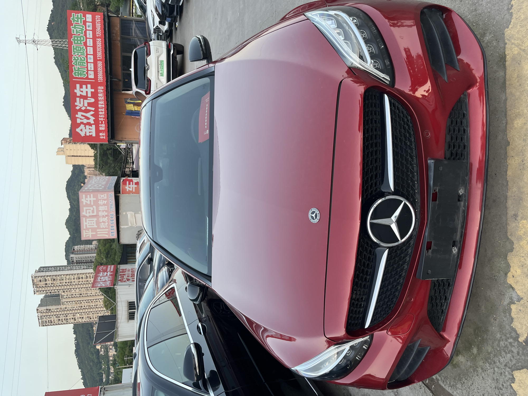 A red Mercedes-Benz car is parked in a lot in Banan, China. In the background are various buildings and signs. One sign reads "金玖汽车" and has phone numbers listed. Other cars surround the Mercedes. The image is taken from a slightly elevated perspective, focusing on the front of the car.