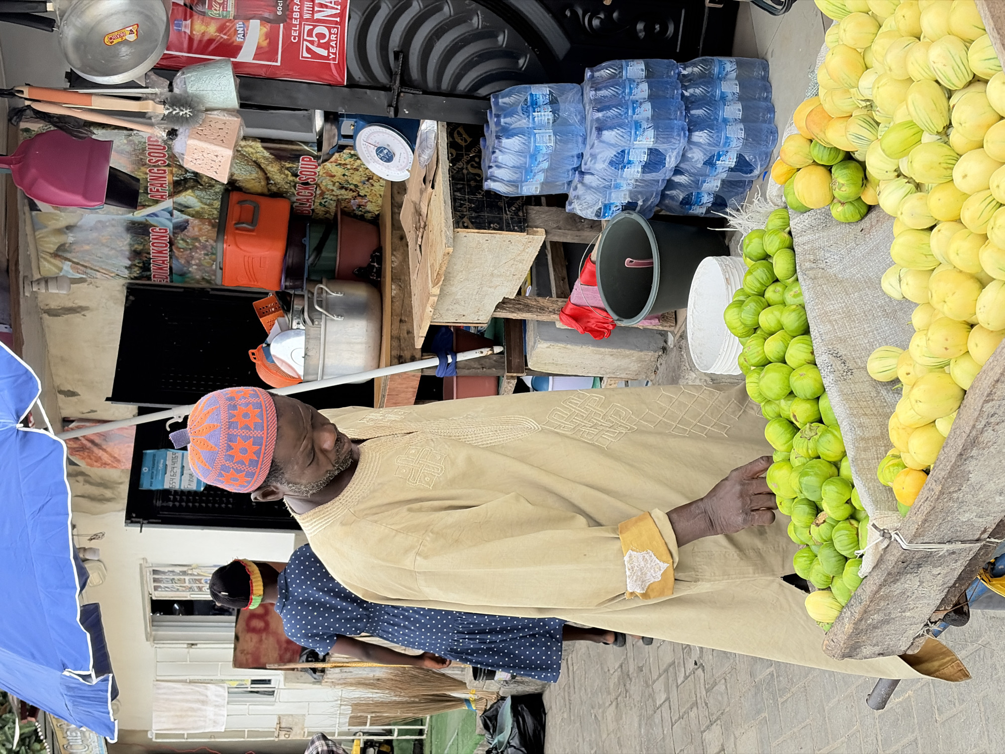 The image depicts a street scene in Igboefon, Nigeria, with a focus on a vendor and his produce. A man, wearing a beige traditional tunic with intricate embroidery and an orange and purple patterned cap, is standing in front of a wooden cart filled with a variety of fruits. He is reaching towards the green fruits in the cart. Behind him, a woman in a dark blue polka-dot dress with a colorful headwrap is walking away from the camera. The background shows a building with windows and a blue awning overhead. Stalls with food items, water bottles, and kitchenware are visible. The ground is paved with interlocking bricks.