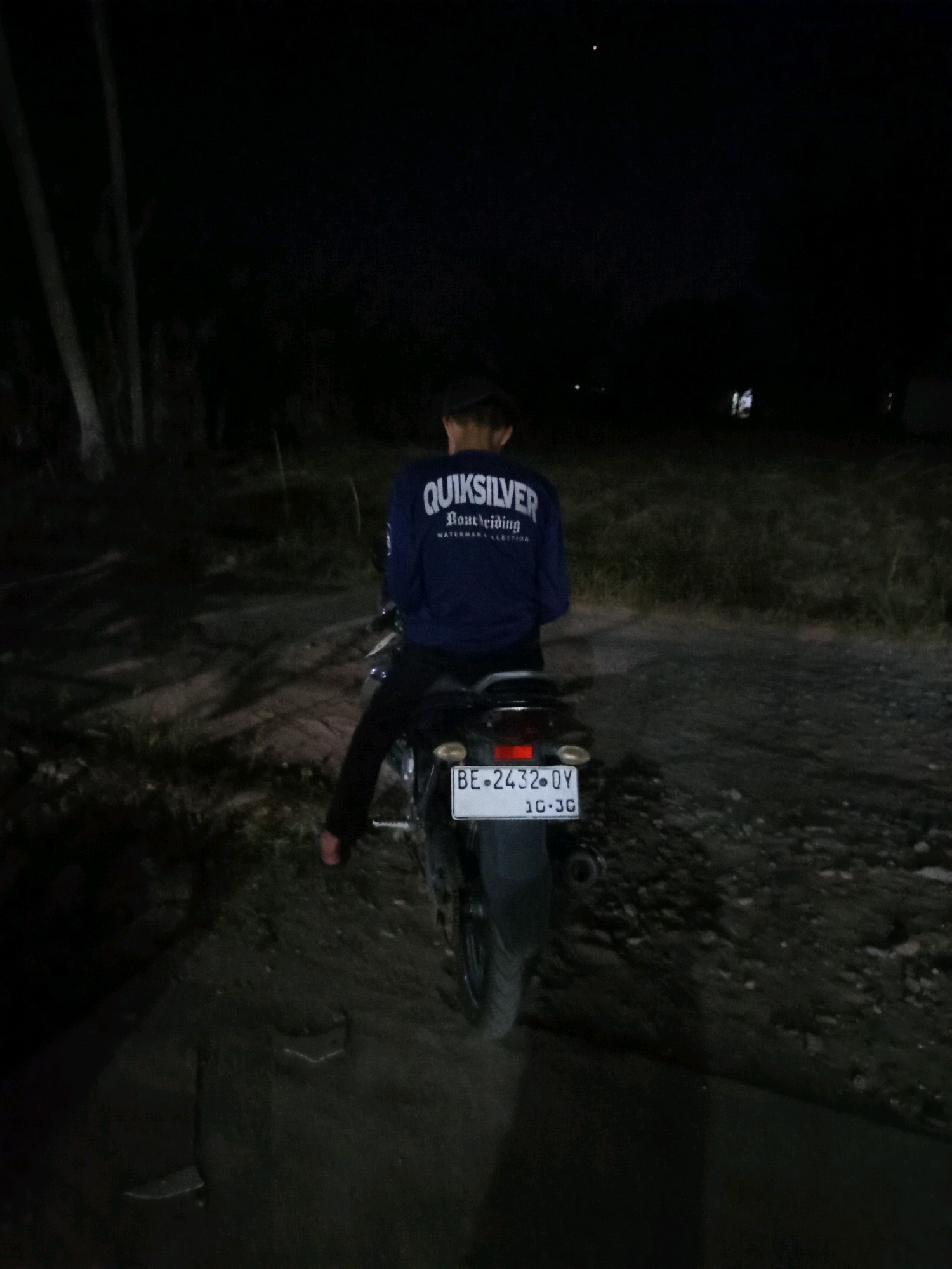 The image depicts a person sitting on a motorcycle at night in what appears to be a rural or semi-rural area. The motorcycle has a license plate that reads "BE 2432 DY" and "10-30". The person is wearing a blue long-sleeved shirt with white lettering that reads "QUIKSILVER". The background is dark, with some trees and vegetation visible. The ground is unpaved, suggesting a dirt or gravel road. There is a faint light in the distance, possibly from a building or streetlights. The overall scene is dimly lit, characteristic of nighttime outdoor conditions.