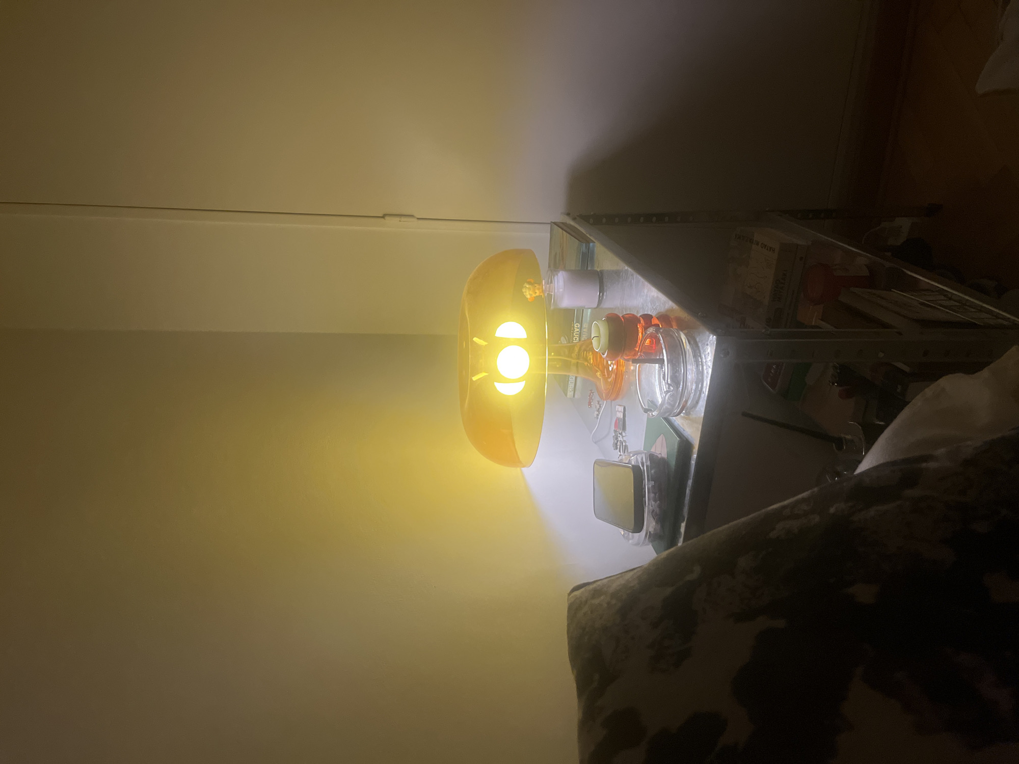 An interior scene, possibly a bedroom or living room, lit by a warm-toned table lamp. The lamp, with an orange mushroom-shaped shade, is placed on a metallic shelf unit. On the shelf next to the lamp are a smartphone, an ashtray containing cigarette butts, a book, and a small candle. The shelf unit also holds several books and other items. In the foreground, a pillow with a cow-print pattern is visible. The wall behind the lamp is lit by its glow, creating a soft ambiance. The image does not contain information that allows for specific location identification within Buenos Aires, Argentina.