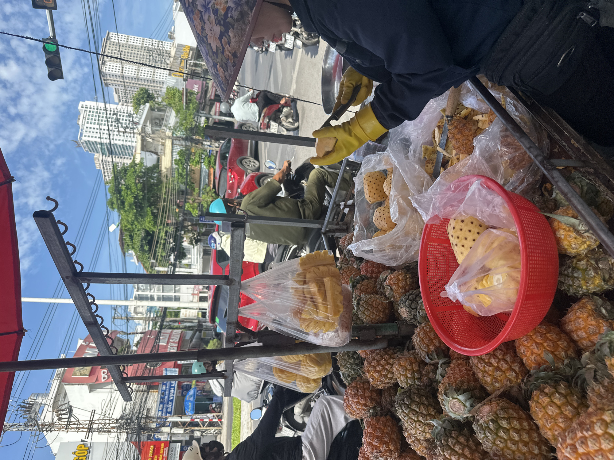 In Bac Nha Trang Ward, Vietnam, a street vendor is seen preparing fruit from a mobile cart. The vendor, wearing yellow gloves and a floral conical hat, is slicing what appears to be pineapple with a knife. The cart is laden with pineapples in various stages of preparation, some whole and others cut. Two large plastic bags filled with sliced pineapple hang from the cart's frame. The background reveals a busy city street with buildings, traffic, and other pedestrians and vehicles. A traffic light displaying green indicates the flow of traffic. The scene is set on a bright, sunny day.