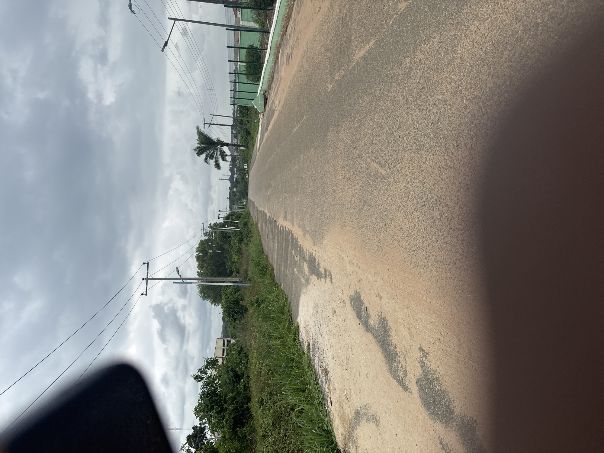 The image depicts a road in Ihiagwa, Nigeria. The road is asphalt and appears to be somewhat worn. Vegetation, including grass and trees, lines the side of the road. Power lines are visible overhead, supported by utility poles. The sky is overcast. In the distance, there is a palm tree and what appears to be a building or structure. A small section of a fence and a sports field is visible along the road to the right. The image is taken at an angle, with the road receding into the distance. A portion of a dark object is visible in the lower left corner of the image.