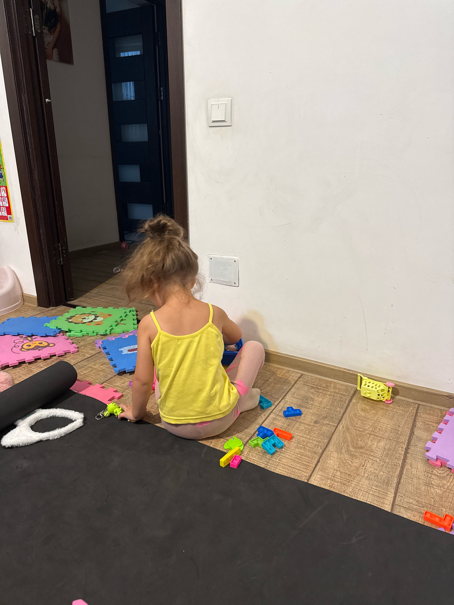A young child with blonde hair in a bun, facing away from the camera, sits on a wooden floor. The child is wearing a yellow tank top and pink leggings. Surrounding the child are colorful foam puzzle mats, some with animal designs. Scattered on the floor are various small toys, including building blocks, a yellow toy shopping cart, and a green frog-shaped keychain. A large black yoga mat is unrolled in the foreground. In the background, a dark wooden door is ajar, revealing a hallway. A light switch is visible on the white wall. The scene appears to be indoors, in what is likely a residential setting in Chișinău, Moldova.