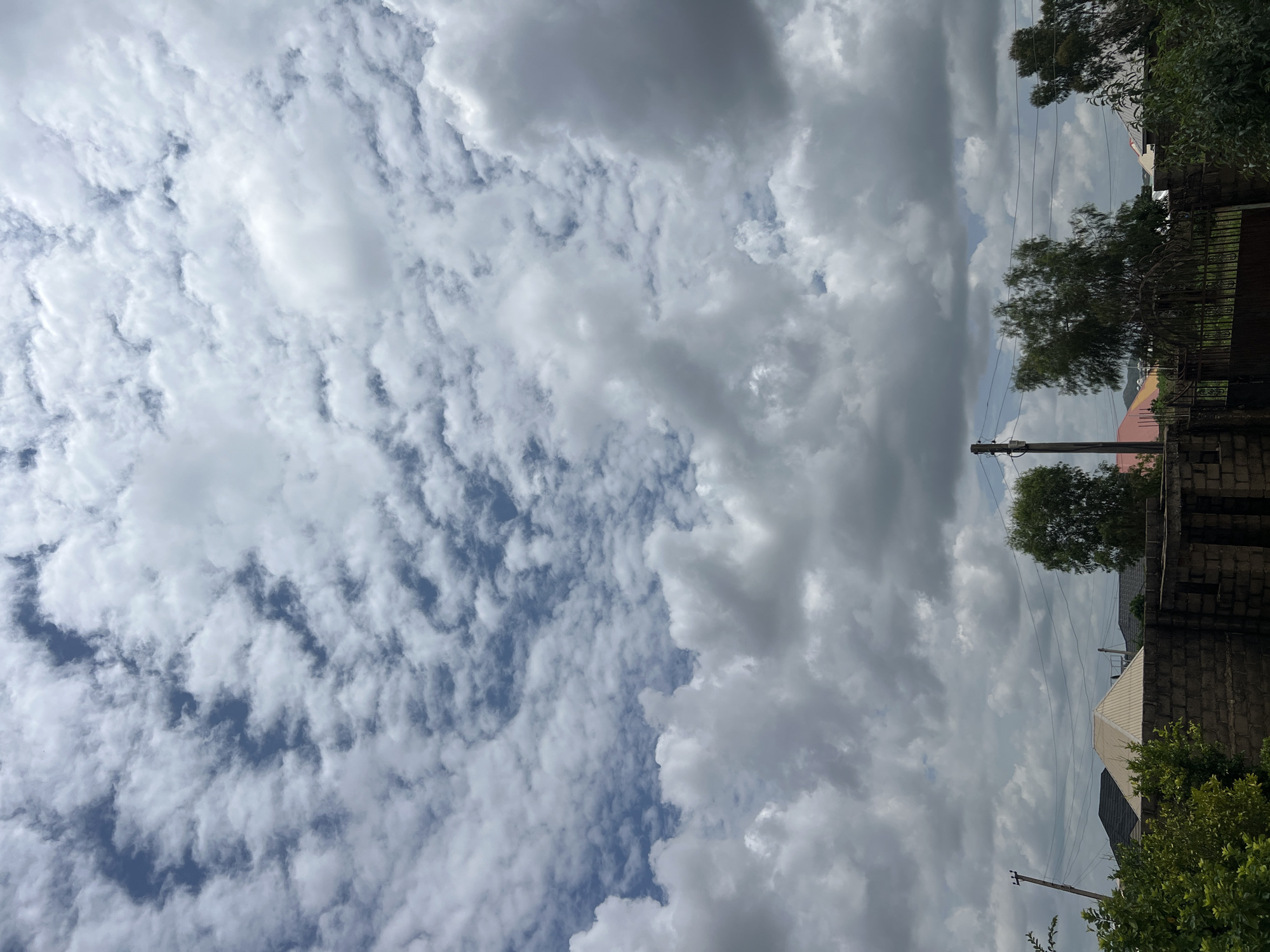 The image depicts a cloudy sky over a residential area in Jos, Nigeria. The sky is filled with a mix of white and gray clouds, with patches of blue sky visible. In the foreground, there is a brick wall and lush green foliage. Behind the wall, several houses with various roof designs are visible. Trees are scattered throughout the scene, some taller than the houses. Electrical poles and wires are also present, indicating the presence of infrastructure. The overall impression is of a typical day in a Nigerian city, with a focus on the sky and the residential environment.