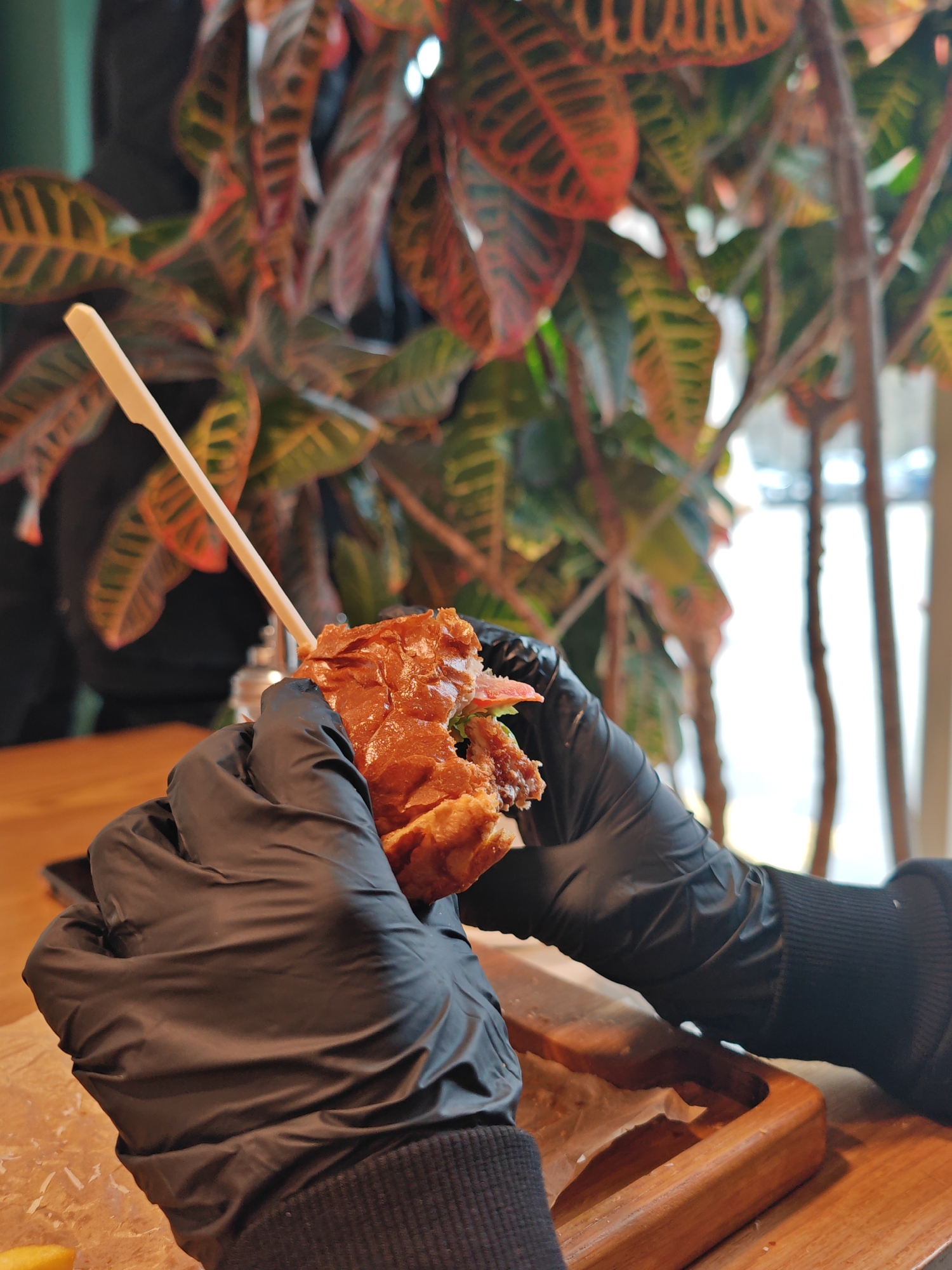 A person wearing black gloves is holding a partly eaten burger in front of a potted plant. A wooden skewer is inserted through the top of the burger. The burger rests on parchment paper on a wooden tray. The scene is taking place in a restaurant in Hrodna, Belarus.