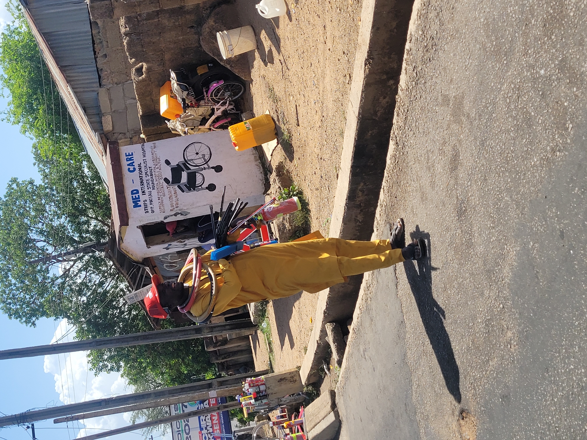 A man in a yellow outfit is walking on the side of a road, carrying a variety of objects on his head and shoulders. He's wearing a red hat and sandals. His shadow is visible on the paved road surface.
Behind him, a building with a sign that reads "MED - CARE STRIPS INTERNATIONAL OPP. PIATEAU STATE SPECIALIST HOSPITAL ROUNDABOUT SUPPLY OF MEDICAL HOSPITAL LABORATORY EQUIPMENT" can be seen. The sign also includes phone numbers. There are other objects near the building, including a bicycle and yellow containers.
In the background, there are trees and a blue sky. In the foreground are utility poles and signs. This scene is located in Jos, Nigeria, as indicated by the sign mentioning Plateu State Specialist Hospital, which is a real hospital in Jos, Nigeria.