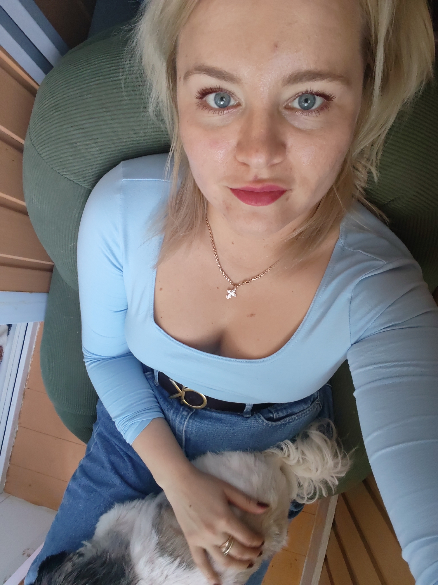 The image shows a close-up of a young woman with blonde hair and blue eyes, wearing a light blue long-sleeved shirt and blue jeans. She is holding a small, fluffy white dog on her lap. The woman is wearing a necklace with a small cross pendant and a belt with a gold buckle. She is sitting in a green armchair. The background suggests an indoor setting with wooden elements and vertical blinds, possibly on a balcony or porch. The lighting appears to be natural daylight. The overall scene is casual and intimate. There is no specific information in the image that would allow for the location to be identified as Izhevsk, Russia.