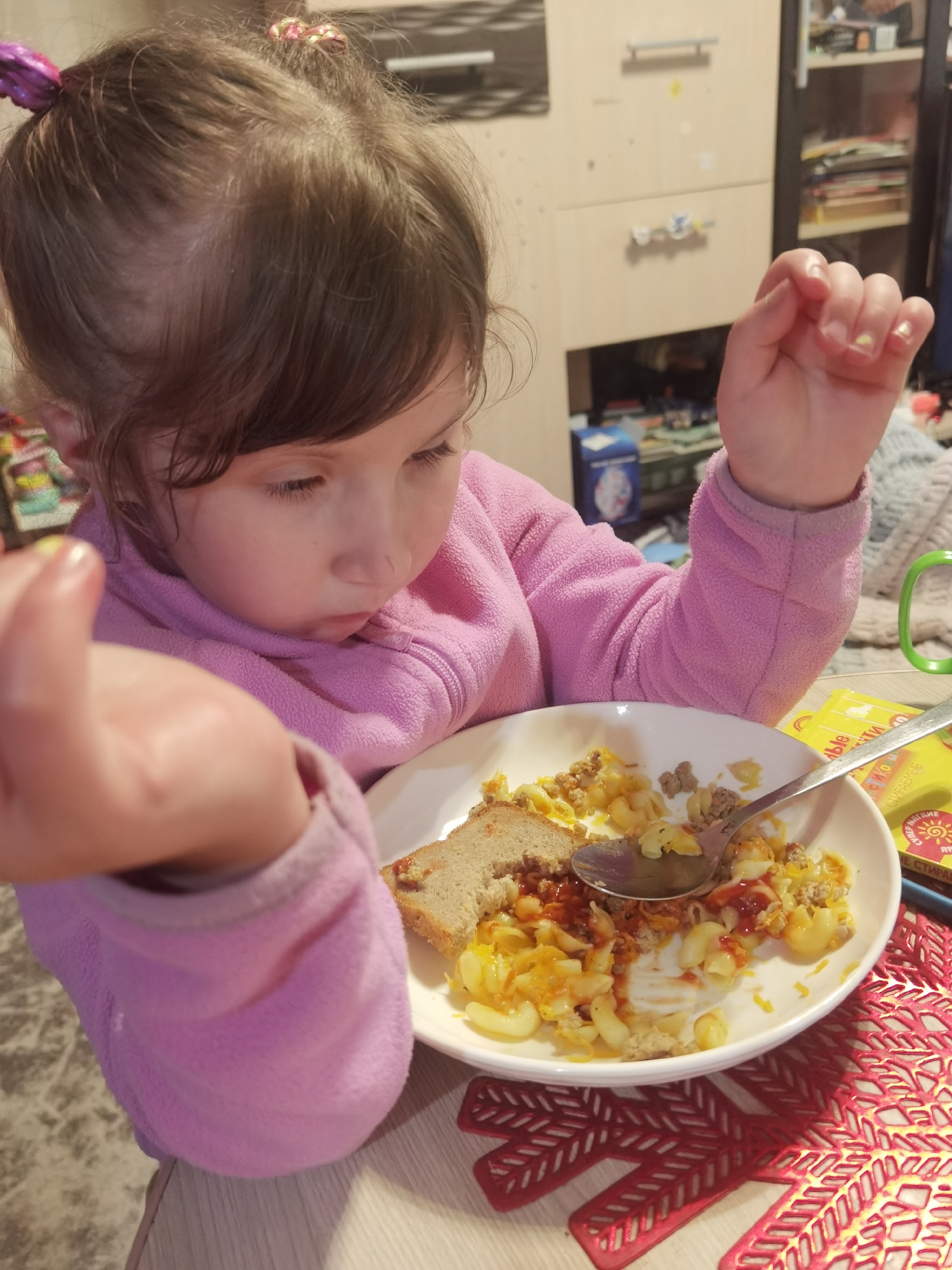 A young female child, estimated between two and five years of age, with medium brown hair styled in two pigtails with pink hair ties, is depicted in a domestic setting within the city of Chaikovsky, Russia. She is wearing a pink fleece jacket. Remnants of light yellow polish are visible on her fingernails. The child is actively engaged in eating, her attention focused on a white circular plate containing elbow macaroni pasta, ground meat, and scrambled egg or cheese, topped with a red condiment resembling ketchup. A slice of brown bread is positioned on the plate. A silver spoon rests within the food. The plate is situated on a light-colored table. A red, open-patterned placemat is beneath the plate. To the right of the plate, a green mug and a yellow item featuring Cyrillic text, including the word 'СТИРАЕМЫЕ' (washable/erasable), are visible. The background consists of light-colored modular cabinetry with various household objects, consistent with an indoor residential environment.
