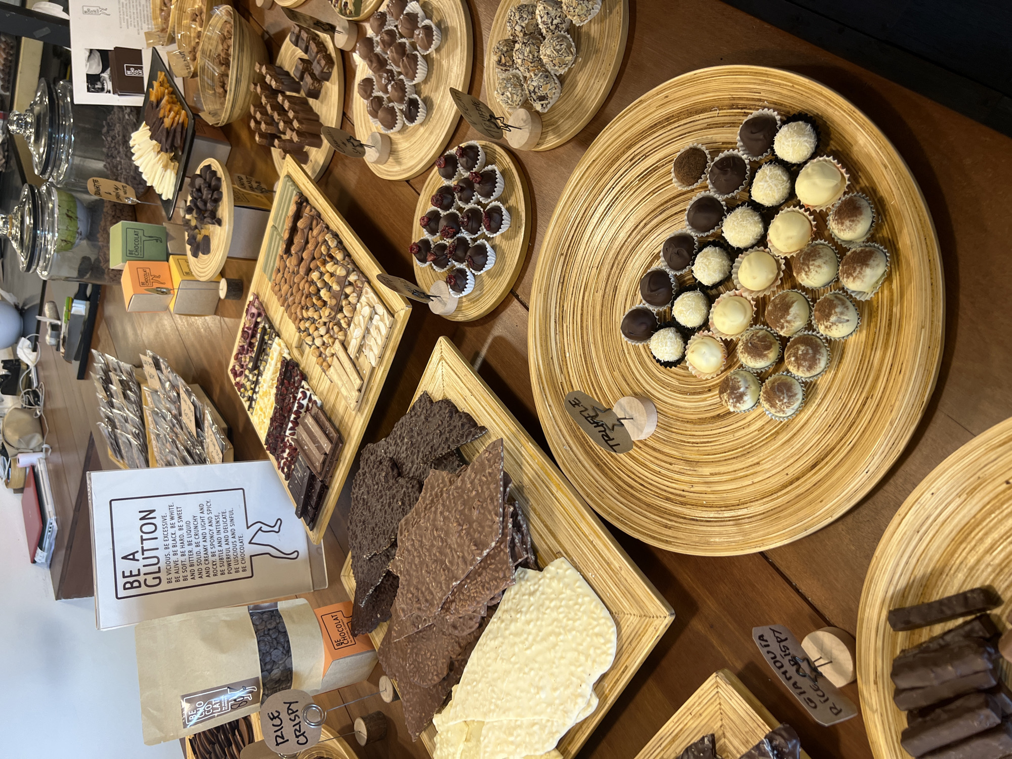 This is a high-angle shot of a chocolate shop in Unknown, Unknown, displaying various chocolate products on a wooden table.
The scene features multiple round and square woven wooden trays holding different kinds of chocolates, including truffles, chocolate barks (white, milk, and dark), and chocolate bars. Some trays have handwritten labels with product names such as "Truffle", "Gianduia Rice Crispy", and "Orangette".
In the background, glass display cases contain more chocolates. Additionally, there's a box labeled "BE A GLUTTON" with text about the characteristics of chocolate. A bag of "Rice Crispy" chocolates is also visible.
The focus is on the variety of chocolate textures and colors, presenting a tempting display for potential customers.