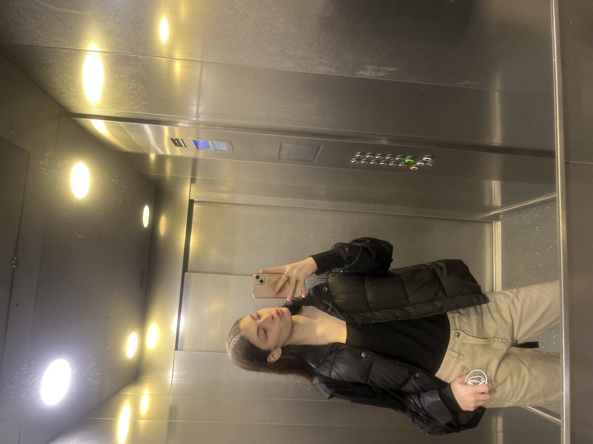 A young woman is taking a selfie in an elevator. The elevator has a silver interior with round, built-in lights on the walls. There are control panels with buttons and digital displays. The woman is wearing a black puffer jacket over a black top and light-colored pants. She is holding her phone in her right hand. She is holding a pair of white earphones in her left hand. It is not possible to determine from the image if the elevator is located in Saint Petersburg, Russia.