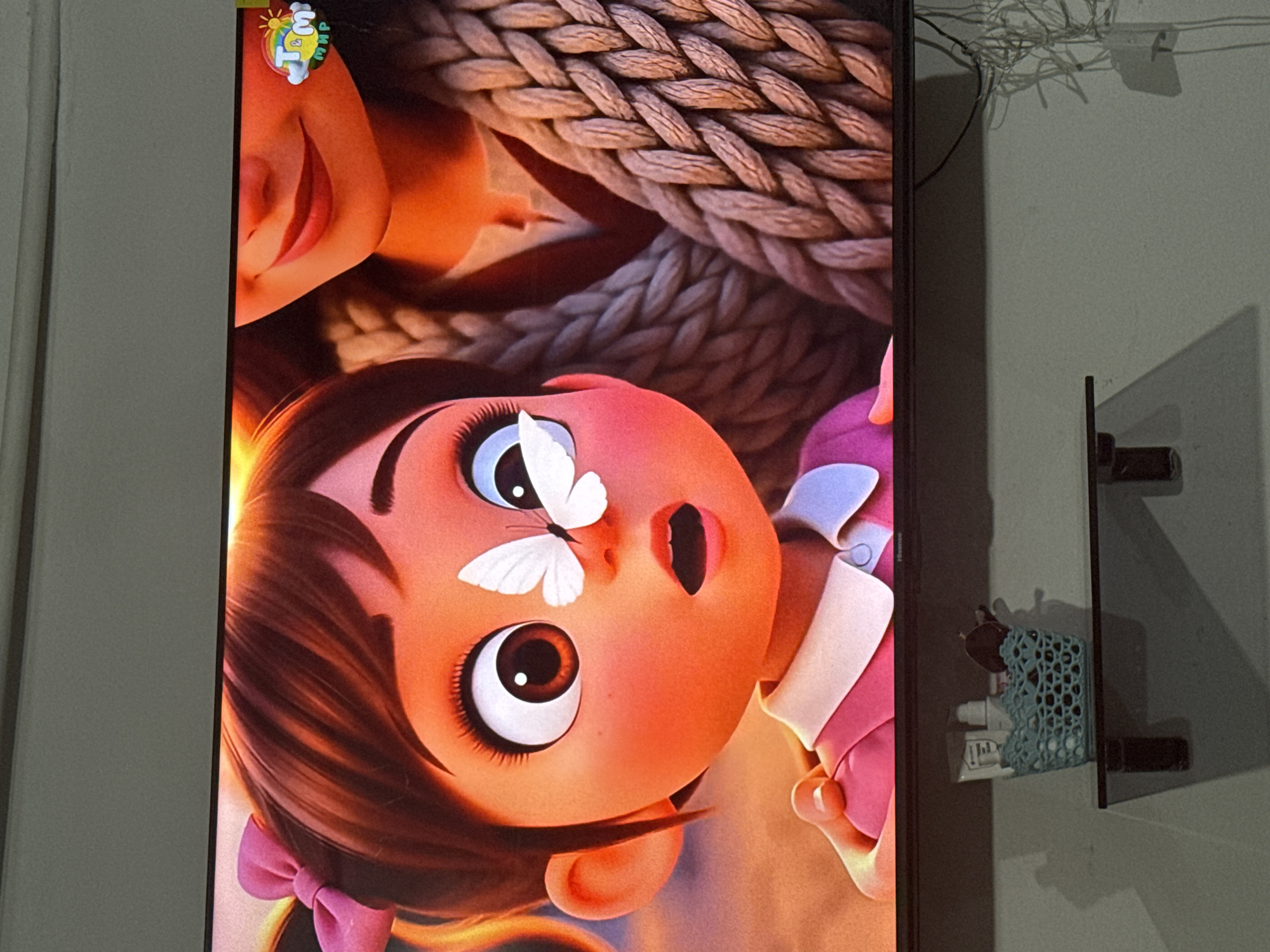 The image shows a close-up of a television screen displaying an animated scene. On the screen, a young girl with large, expressive eyes and a pink bow in her hair has a small white butterfly perched on her nose. She is wearing a pink dress with a white collar. Beside her, a portion of another person's face, likely an adult, is visible, with red lips and a braided, textured garment. In the upper right corner of the screen, a colorful logo featuring a rainbow and text is displayed. Below the television, a small light blue crocheted basket sits on a shelf, containing several bottles and dark-colored objects, possibly sunglasses. The background appears to be a wall. The location is specified as Chardara, Uzbekistan, however, this detail is not visually evident from the image itself.
