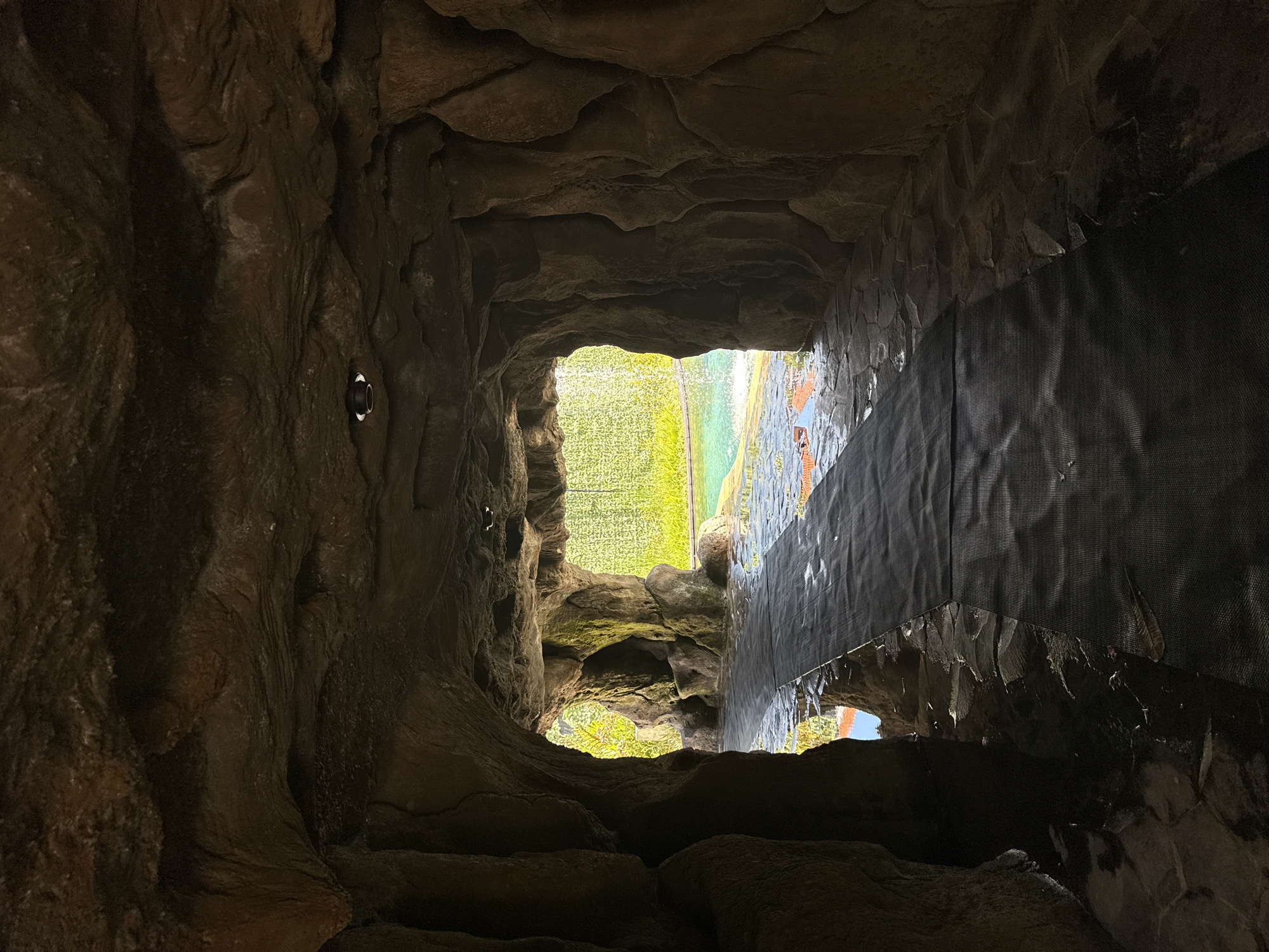 View looking outward from within a dark, simulated rock structure. The structure’s walls are uneven and textured, resembling the inside of a cave. On the right, a black, mesh-like material is partially covering the constructed rock. The opening reveals a bright, green, grassy area, beyond which are hints of blue sky. Nusa Dua, Indonesia.