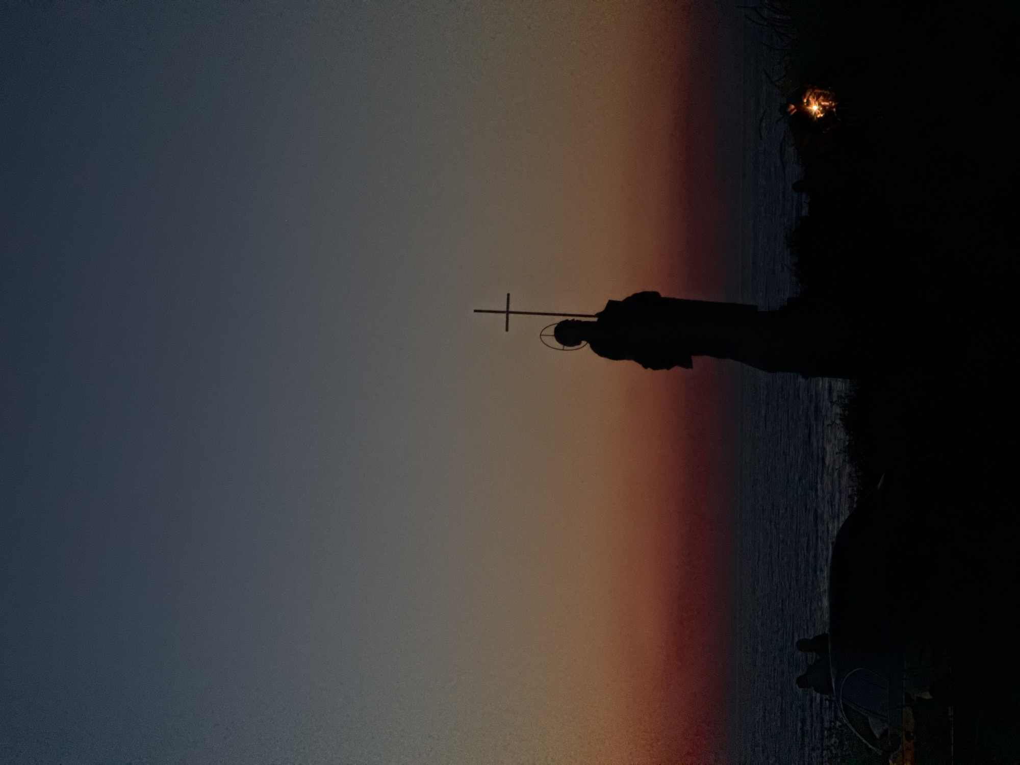 A silhouette of a statue holding a cross stands on a hill overlooking the ocean at sunset. The sky is a gradient of deep blue to orange and red. In the foreground, a car and a few figures are also silhouetted against the darkening landscape. A small fire or light source is visible to the right of the statue.