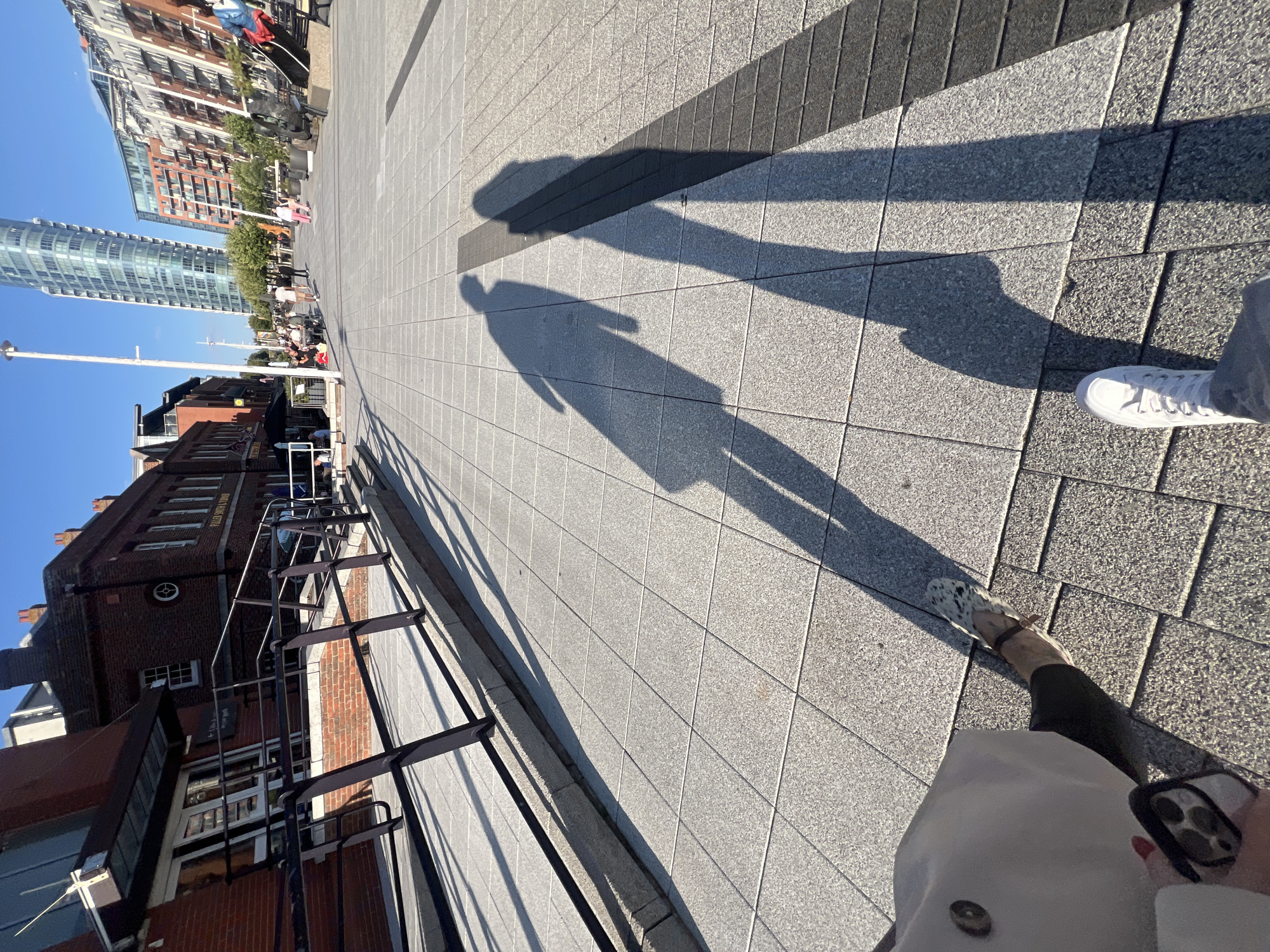 This image depicts a sunny day in Portsmouth, United Kingdom, likely in the modern Gunwharf Quays area, characterized by a mix of historic brick buildings and contemporary high-rise architecture. In the foreground, the shadows of two individuals are cast upon a patterned paved walkway. One person's feet are visible, wearing white sneakers and dark trousers, holding a phone. The other person is wearing distinctive patterned flat shoes and dark trousers.
The walkway is made of large, light grey square tiles with dark grey rectangular tiles forming a distinct strip. To the left, a low brick wall with a metal railing runs along a sloped ramp, suggesting accessibility features. Behind the railing, a red brick building with large glass doors and windows is visible, labeled "Fuller Smith & Turner." Further down the street, modern buildings with glass facades and balconies rise, one notably a tall, slender skyscraper with a curved top. Several people are scattered throughout the scene, walking or seated at outdoor tables, enjoying the pleasant weather. The sky is a clear, vibrant blue.