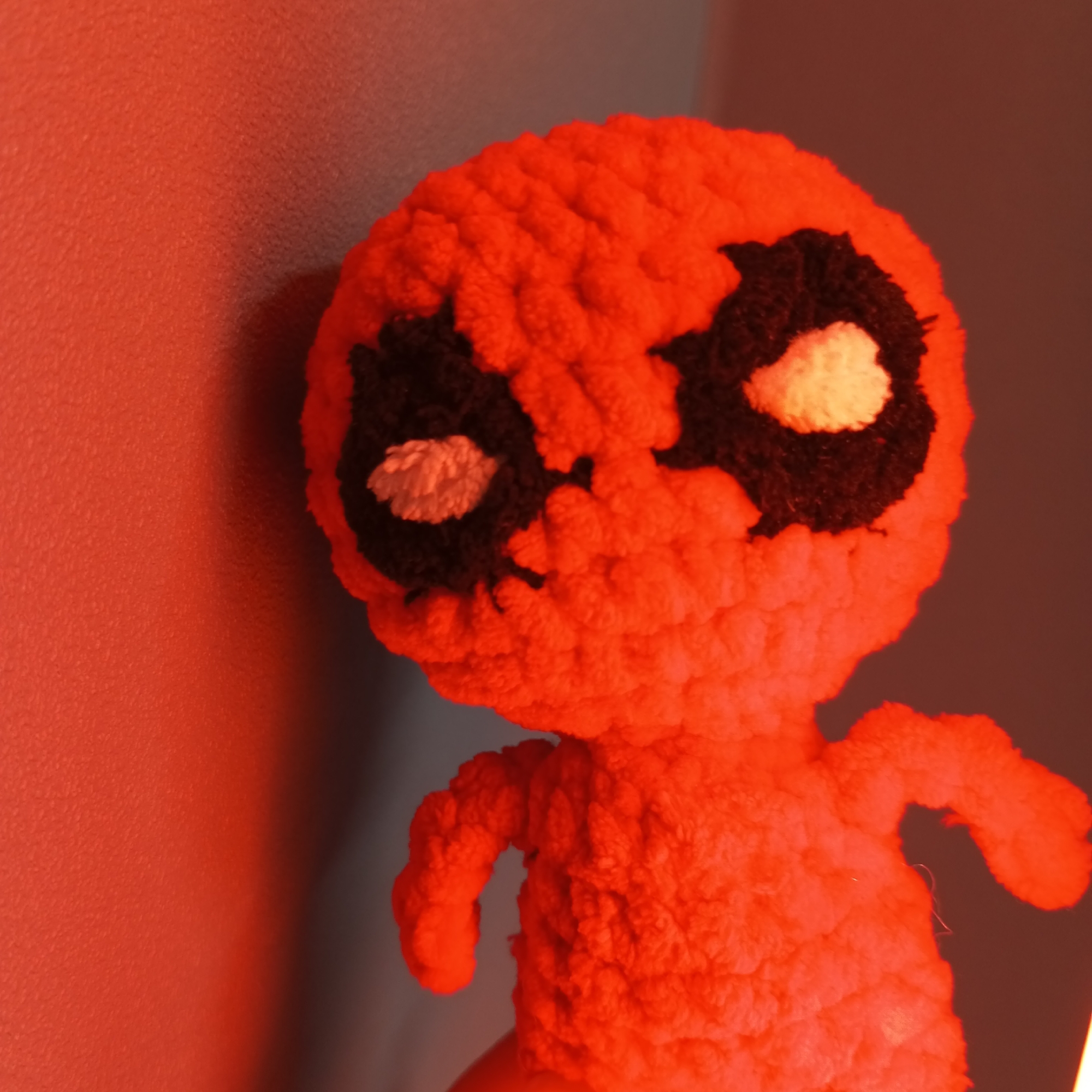 A handmade, plush Deadpool doll with black eye patches and white eye highlights stands against a red wall. The lighting is red, and the doll is positioned on a surface. The location is not discernible from the image, and therefore Pervomaysky, Russia cannot be verified.