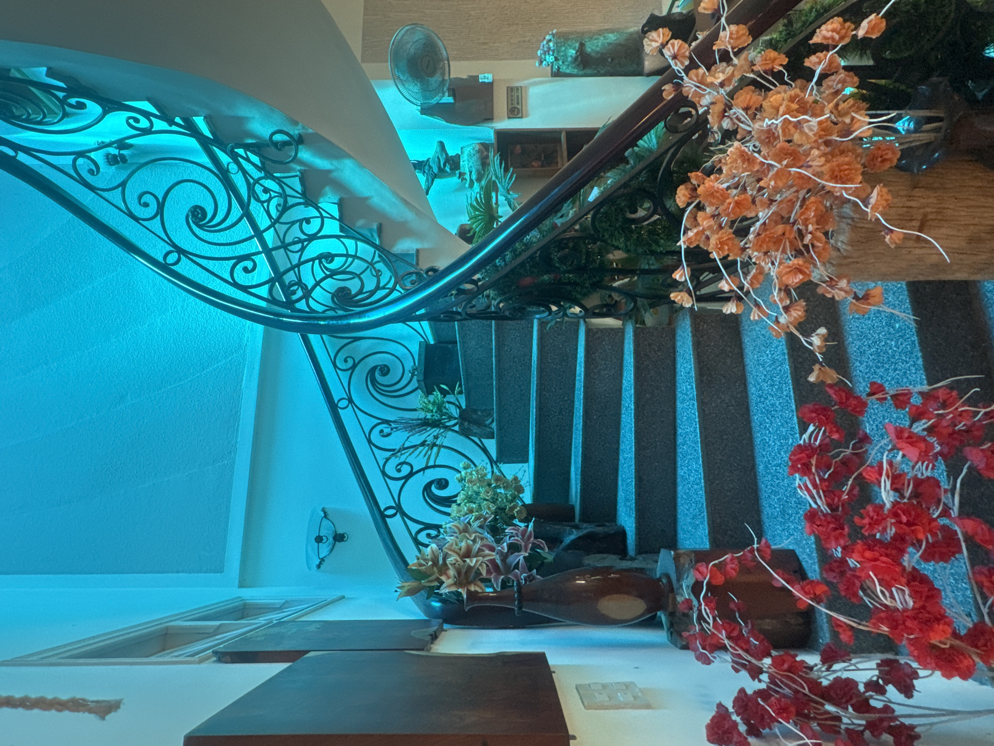 The image depicts a spiral staircase in what appears to be a hotel or residential building, illuminated by blue lighting. The staircase has a dark metal railing with an intricate, swirling design. The steps are made of a speckled gray material. Several decorative elements are placed around the base of the staircase. On the left, a tall, dark brown vase with a lighter oval design stands next to a wooden structure, with red artificial flowers in the foreground. On the right, a wooden log holds a bouquet of orange artificial flowers. In the background, a small fan is visible on a shelf, along with other decorative items and plants. The overall impression is one of a decorated interior space, possibly in a tropical location given the lush greenery and floral arrangements. Without further context, the specific location within Bac Nha Trang Ward, Vietnam, cannot be determined from the visual information alone.