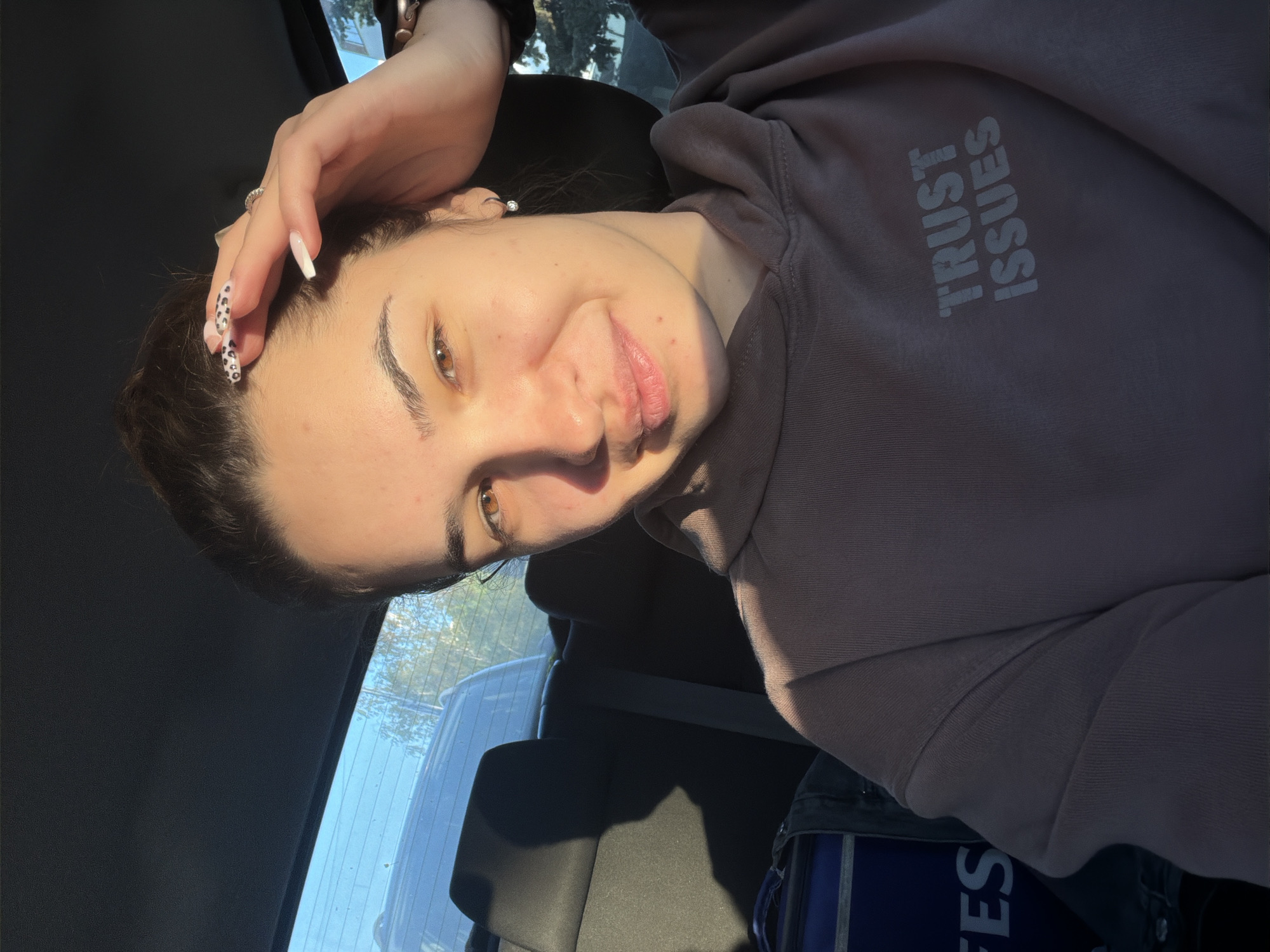 A woman is taking a selfie inside a vehicle. She is wearing a purple hoodie with the words "TRUST ISSUES" printed on it. Her hand is resting on her head, showing long, patterned nails. A glimpse of the outside through the car window reveals foliage and what appears to be other vehicles. The location is most likely not Sofia, Bulgaria as there is no identifying feature of the city present in the image.