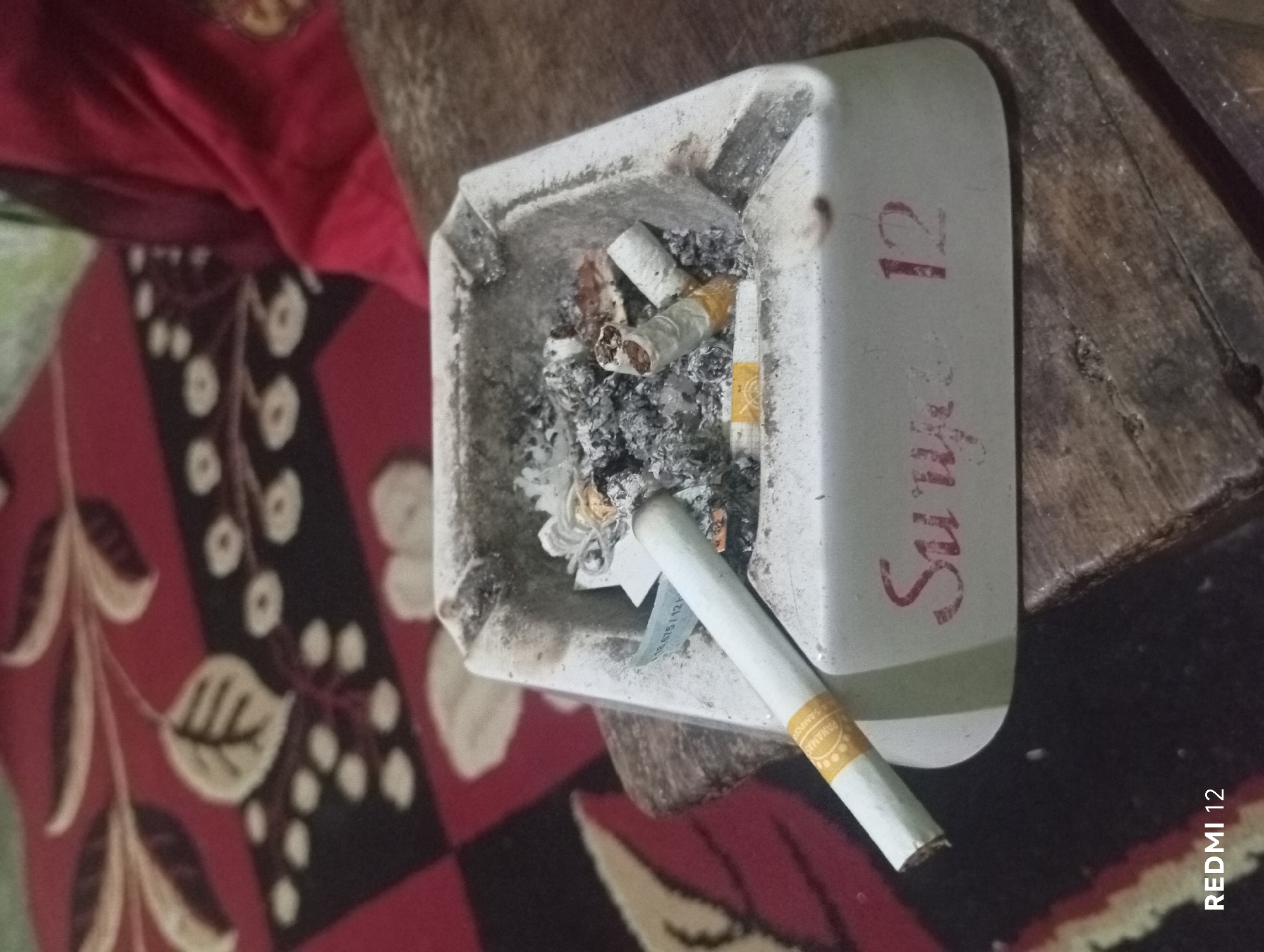 An ashtray filled with cigarette butts and ash sits on a wooden surface. A lit cigarette rests on the edge of the ashtray, with a partially smoked cigarette inside. The ashtray has red lettering on it that reads "Surya 12". In the background, a patterned fabric is visible, with shades of red, black, and white. The scene appears to be indoors.