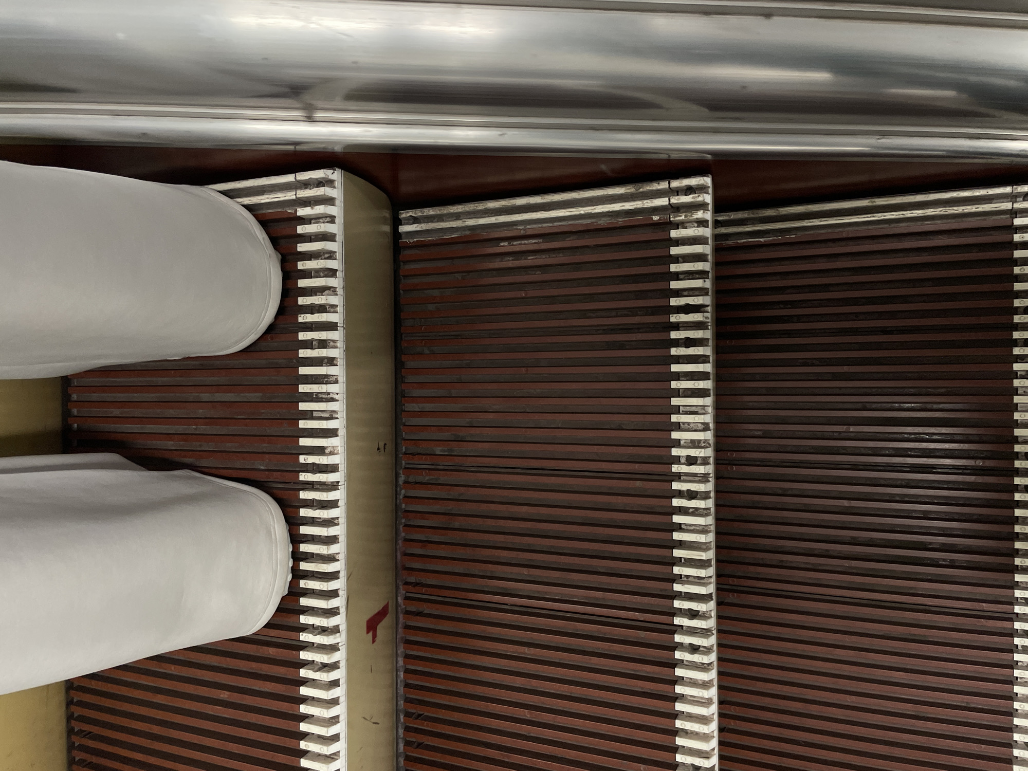 The image shows a close-up, top-down view of an escalator. Two white, padded handrails dominate the left side of the frame, curving downwards. The escalator steps are made of dark brown material with thin, white safety strips along their edges. The steps are ribbed, creating a textured surface. The overall scene suggests an indoor environment, possibly a public transportation hub or a large building. There is no visible text or indication of time of day or weather. The image is oriented vertically, suggesting it was taken while the camera was held upright.