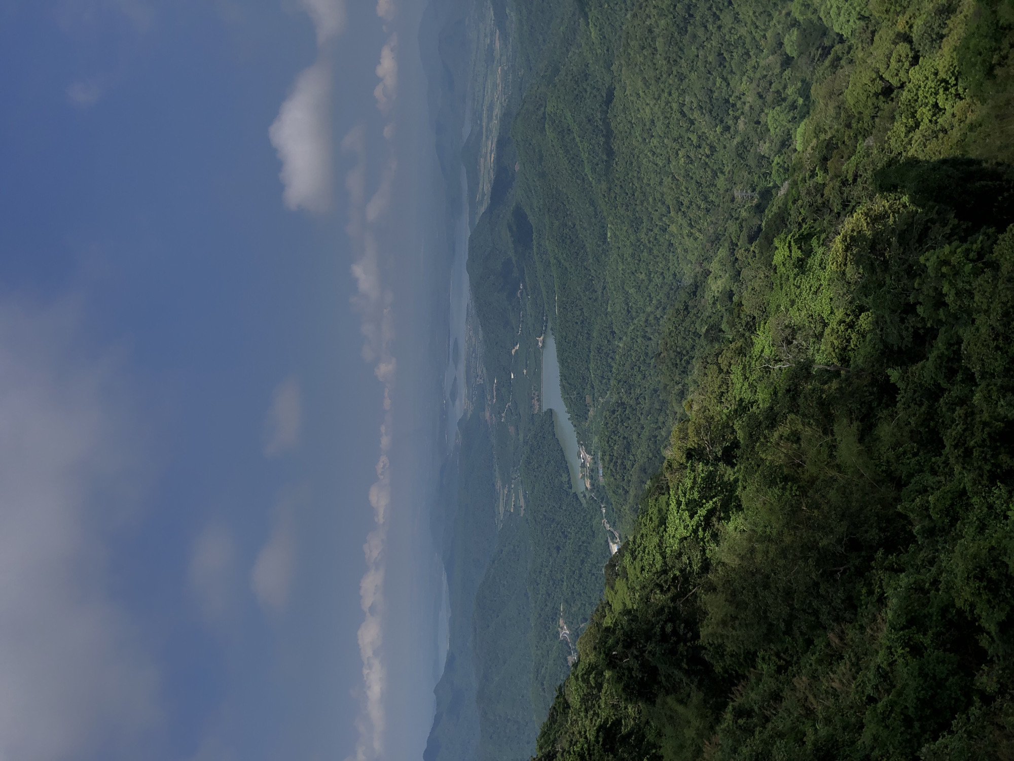 This is a high-angle, wide shot of a lush, mountainous landscape with a body of water in the distance, likely taken from a viewpoint in Nam Hải, Vietnam. The foreground is dominated by a steep, verdant mountainside covered in dense green foliage, with some trees casting shadows. The midground reveals rolling hills covered in a similar thick forest. In the distance, a winding river or inlet, with some structures visible along its banks, flows through the valley. Beyond the immediate hills, a series of progressively more distant mountain ranges fade into a hazy, atmospheric perspective under a blue sky dotted with scattered white clouds. The scene suggests a natural, undeveloped area with evidence of some human habitation and infrastructure, such as roads and buildings, integrated into the landscape.