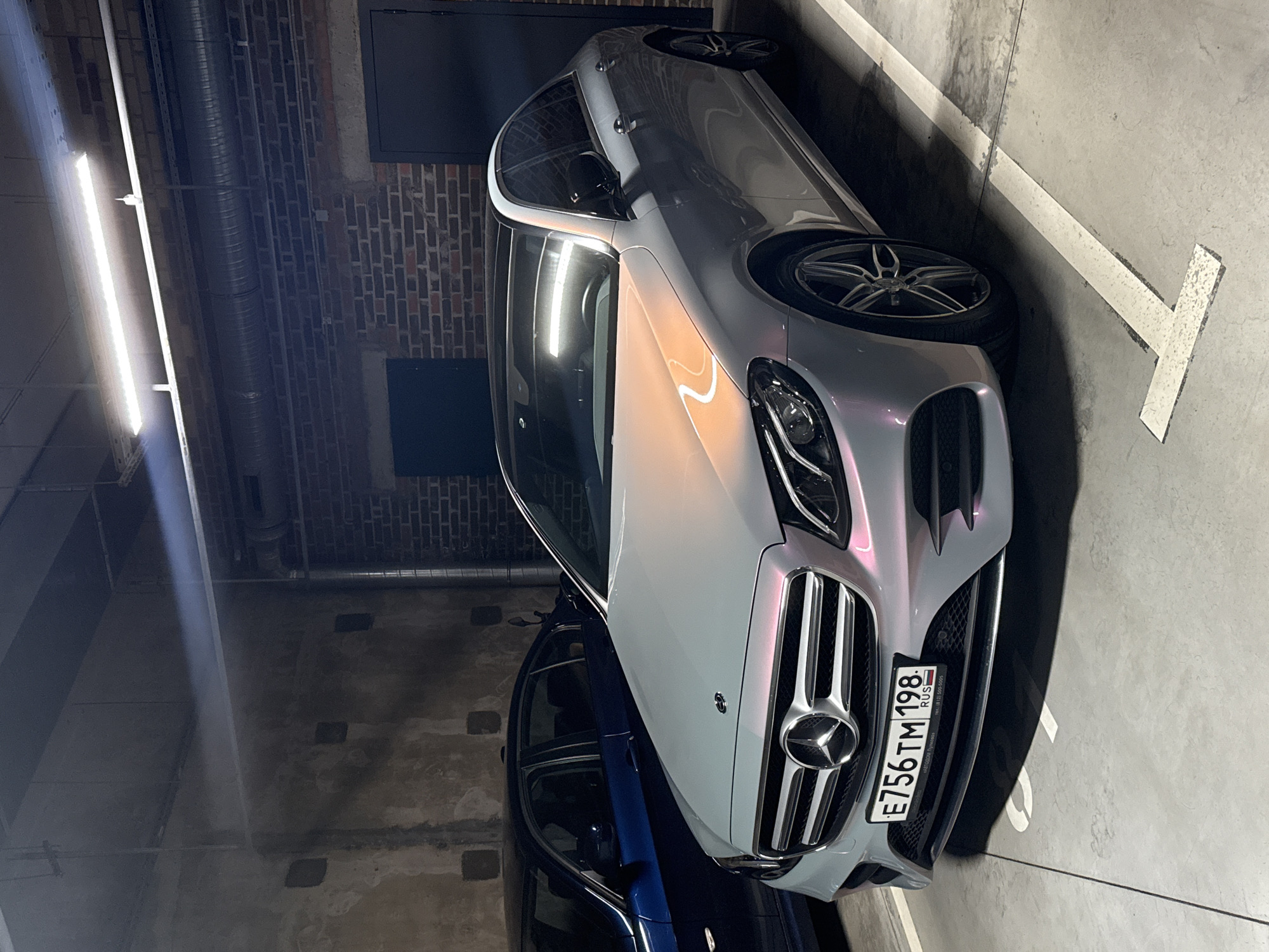 A silver Mercedes-Benz sedan, likely an E-Class, is parked in an indoor parking garage. The car's paint exhibits a multi-tonal, iridescent quality, reflecting hues of pink, yellow, and blue from the overhead artificial lighting. The vehicle features a prominent chrome grille with the Mercedes-Benz star logo and multi-spoke alloy wheels.
The front license plate is clearly visible, displaying "E756TM 198 RUS". The "198" region code indicates Saint Petersburg, Russia, aligning with the provided location context. Smaller text beneath the main characters on the plate includes a phone number: "(8 813) 590-50-66" and "РОССИЯ" (Russia).
The setting is a dimly lit concrete parking structure. Fluorescent strip lights illuminate the area, casting strong reflections on the car's surface. In the background, sections of a brick wall and exposed metal ventilation pipes or conduits are visible overhead. A dark blue car is partially visible parked next to the Mercedes-Benz. White parking lines, including a distinctive "T" or arrow-like marking, are painted on the concrete floor. The overall impression is an underground or multi-story parking facility.
