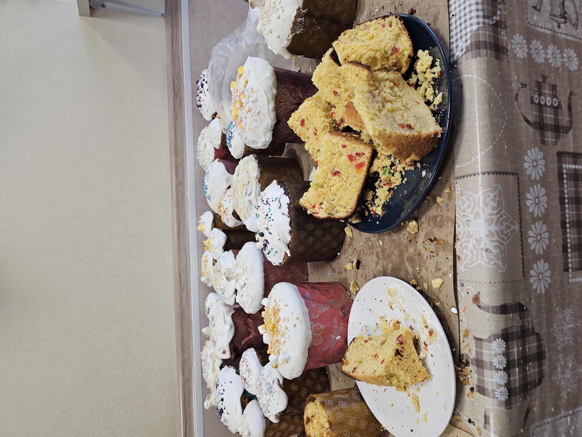 The image depicts a table covered with a patterned tablecloth with images of teapots and flowers. Several Easter breads (kulichi) with white icing and sprinkles are arranged on the table. Some of the breads are whole, while others are cut and served on blue and white plates. Crumbs are scattered around the plates and breads. The image appears to have been taken indoors, possibly in a home setting, as evidenced by the adjacent wall. There is no information to determine if the location is in the city of Khimki, Russia.