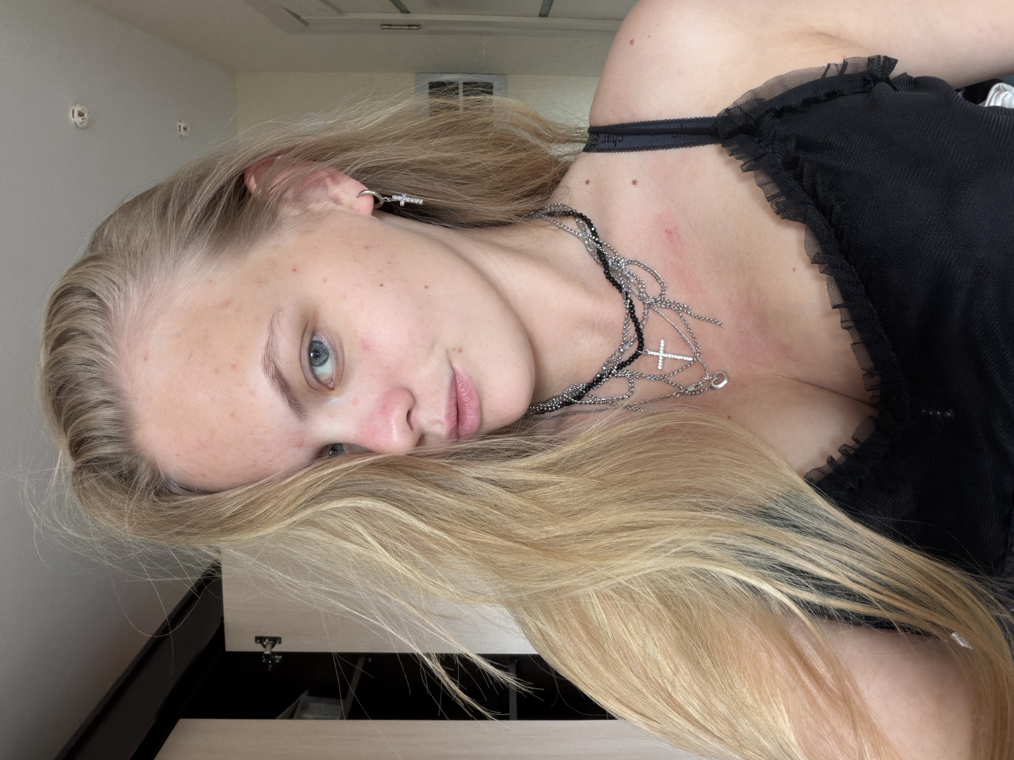 A close-up portrait shows a young woman with long, straight blonde hair and blue eyes, looking directly at the camera with a neutral expression. The image is rotated approximately 90 degrees clockwise, placing her head on the right side of the frame. Her blonde hair cascades around her face and over her shoulders and chest.
She wears a black top with a delicate, ruffled mesh trim around the neckline or straps, and a black bra strap is visible. Multiple layered necklaces adorn her neck, prominently featuring a silver-toned chain with a large cross pendant. A matching silver-toned earring with a small cross design is visible in her left ear. Her fair skin shows some red blemishes on her forehead, cheek, and chest.
The setting appears to be indoors, with soft, natural lighting indicating daytime. The background consists of light-colored walls and a section of a light-colored furniture piece with a dark trim. Faint, indistinct shapes are visible on the wall in the distance. No specific activity beyond posing for the photograph is depicted. A faint, unreadable pattern or text is visible on a black elastic band near her right shoulder. The image provides no specific visual cues to its Izhevsk, Russia, context.