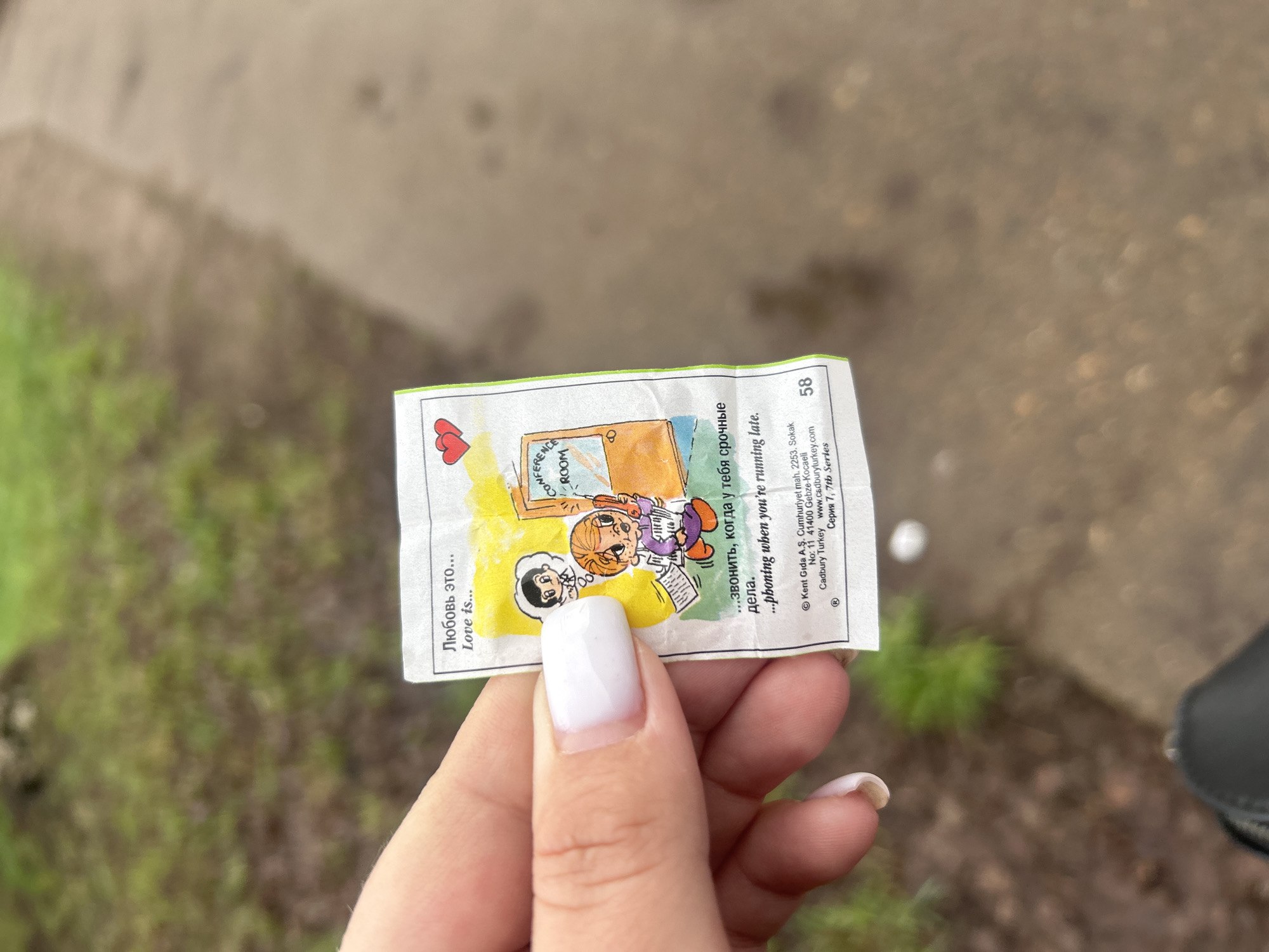 A close-up shot captures a person's hand, adorned with white nail polish, holding a small, colorful "Love is..." chewing gum wrapper. The wrapper features a classic cartoon of a man with black hair and a woman with orange hair, wearing a purple top and orange trousers, busily talking on a phone while holding a book, with a "CONFERENCE ROOM" sign visible in the background. A small red heart symbol is also printed on the wrapper.
The text on the wrapper, in both Russian and English, reads: "Любовь это... Love is... ...звонить, когда у тебя срочные дела. ...phoning when you're running late." Below this, smaller print identifies the manufacturer and origin: "® Kent Gıda A.Ş. Cumhuriyet mah. 2253. Sokak No: 11 41400 Gebze-Kocaeli Cadbury Turkey www.cadburyturkey.com Серия 7, 7th Series," along with the number "58."
The background is out of focus, showing a blurry outdoor setting with patches of light brown ground or a path and hints of green grass or foliage. Part of a dark shoe or boot is visible in the lower right corner. The image was taken in Chaikovsky, Russia.