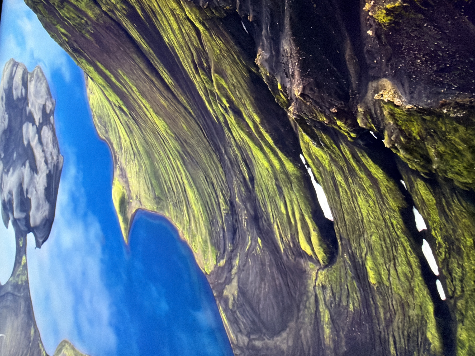 The image shows an aerial view of a stark, mountainous landscape with a vibrant blue body of water. The terrain is characterized by steep, moss-covered slopes that exhibit a textured, striated pattern. Patches of dark, rocky ground are interspersed with the bright green moss, particularly on the higher elevations and the exposed rock faces. The body of water, likely a lake or sea, is a deep blue and reflects the sky, with some clouds visible on its surface. The overall impression is one of rugged natural beauty, with dramatic contrasts between the water, the verdant moss, and the dark earth. There is no indication of any urban environment or any connection to the city of Kyiv, Ukraine.