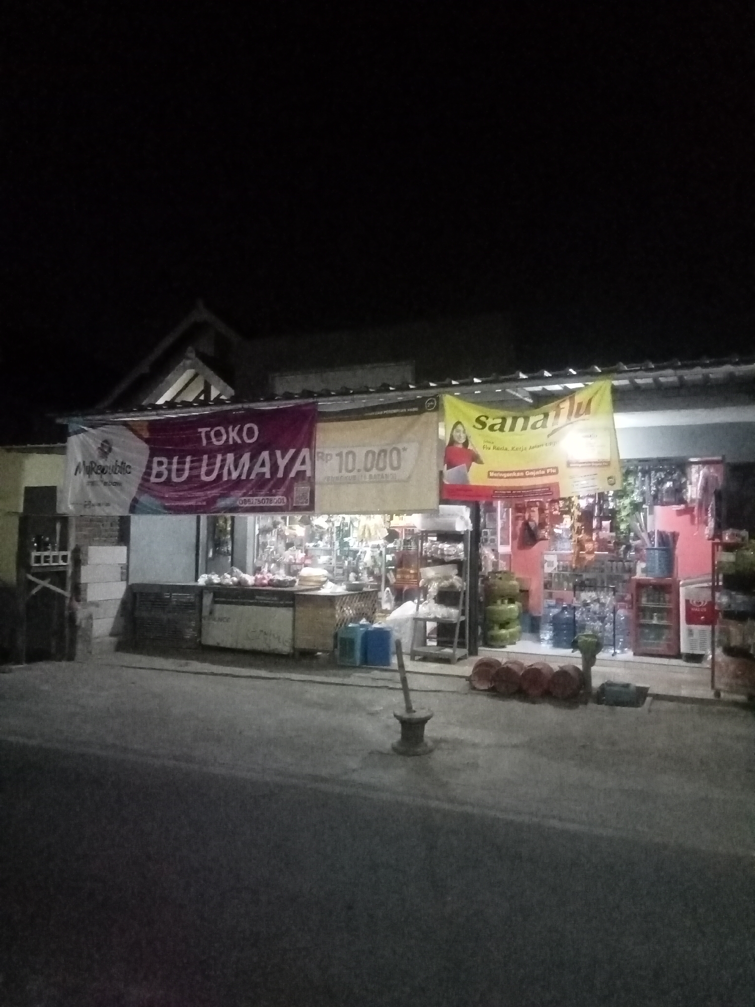The image depicts a street-level view at night, possibly in the city of Malang, Indonesia, in front of a small shop. The shop appears to be a general store or warung, indicated by the shelves stocked with various goods and bottles of drinks visible through the open storefront. Two prominent banners hang above the shop. The banner on the left is white and features a logo with the text "MyRepublic". Next to it, a larger, dark pink banner reads "TOKO BU UMAMA" and also displays "Rp 10.000". A yellow banner to the right advertises "sanaflu" with an image of a woman and text in Indonesian related to flu relief. In front of the shop, several green gas cylinders are stacked, along with red and white circular objects that could be drums or large containers. A person, partially visible and wearing a reddish garment, is seen inside the shop. The foreground shows a paved street with a rough texture, and a dark sky is visible above. The scene is illuminated by artificial light, creating shadows and highlighting the shop's facade.