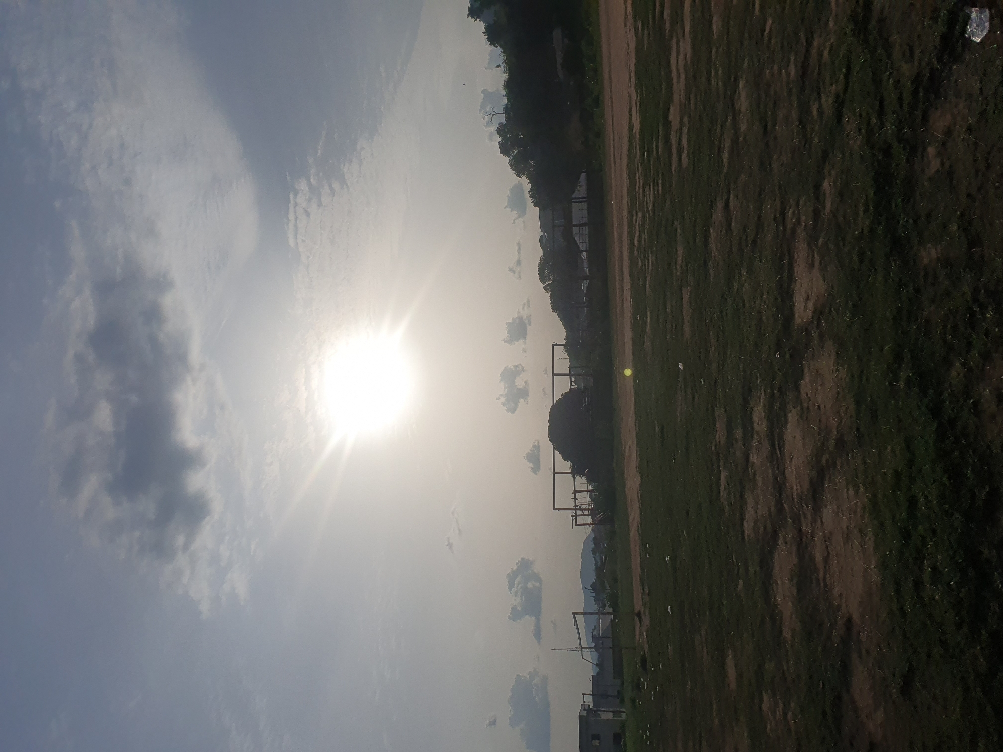 The image shows a landscape in Lokoja, Nigeria, under a bright, hazy sky. The sun is prominently positioned, shining through a layer of clouds and haze. The foreground consists of a grassy and dirt field, typical of a sports ground or open area. In the middle ground, there are structures that seem to be goalposts or fencing, possibly marking a boundary of the field. Further in the background, there is vegetation, possibly trees or bushes, along with building structures that are typical of an urban environment. The horizon is somewhat blurred due to the haze and the camera angle. A piece of trash is visible in the far upper right corner. The image is rotated counterclockwise.