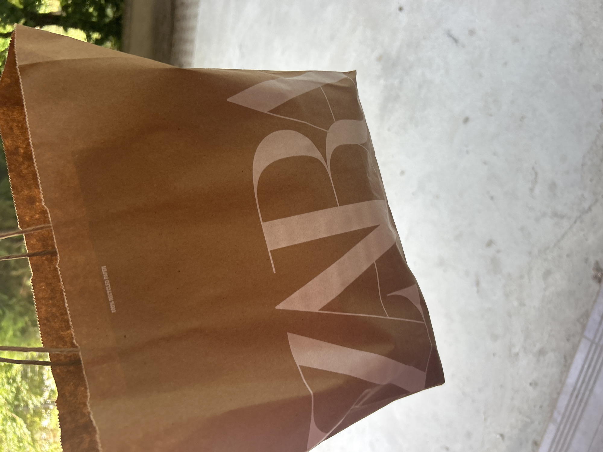 A brown paper shopping bag with large, light-colored lettering that reads "ZARA" is the primary subject of this media file. The bag is positioned diagonally, with its top edge and handles visible on the left side, suggesting it is being held or has just been placed down. The texture of the paper is visible, along with a serrated edge along the top. The bag has "100% RECYCLED PAPER" printed vertically along its side in small text. The background is a light-colored, mottled surface, possibly concrete or stone, which appears to be outdoors. Green foliage is visible in the upper left corner, indicating an outdoor setting with trees or other plants. The lighting suggests it could be daytime. The image is cropped closely on the bag and the immediate background. There are no people or animals visible in the frame. The overall mood is neutral, focusing on the object itself.