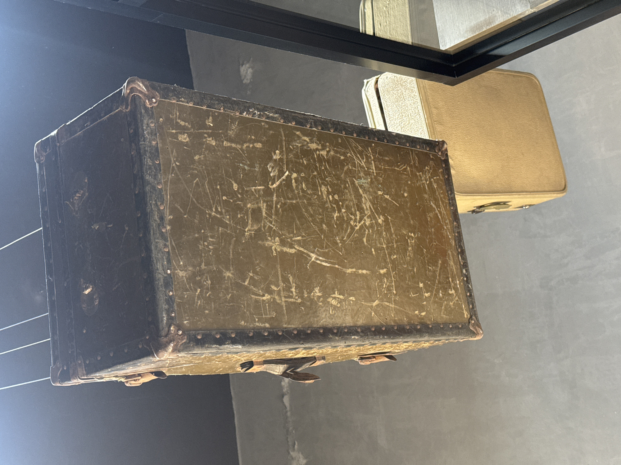 The image displays a close-up view of vintage luggage, likely part of an indoor art installation or exhibit. The primary subject is a large, rectangular, olive-green or dark brown trunk positioned prominently in the foreground, oriented diagonally. This trunk shows significant wear, with numerous white scratches and scuff marks across its surface, indicating age and extensive use. Its edges are reinforced with dark material, possibly metal or leather, secured with visible rivets, and it features metal corner protectors, some showing rust or patina. Two metal latches or clasps are visible on its underside. The trunk appears to be suspended by thin, white wires or strings, which are faintly visible on the left side.
In the background, positioned above and to the right of the main trunk, are several lighter-colored, beige or tan suitcases. One rigid, vintage-style suitcase is clearly visible, also appearing to be suspended or placed on a high surface. A black, reflective beam or frame runs horizontally across the upper part of the image, contributing to the structural elements of the display. The background consists of a plain, light gray wall, likely concrete, suggesting an exhibition space.
No direct activity or people are visible. The scene captures a static display designed to highlight the historical or aesthetic qualities of vintage travel items. The lighting appears to be artificial, illuminating the textures and details of the luggage. There is no visible text in the image. The arrangement suggests a curated environment, possibly in Apetatitlán, Mexico, where these items are presented for viewing. The overall impression is one of nostalgia and the passage of time, encapsulated in the worn luggage.